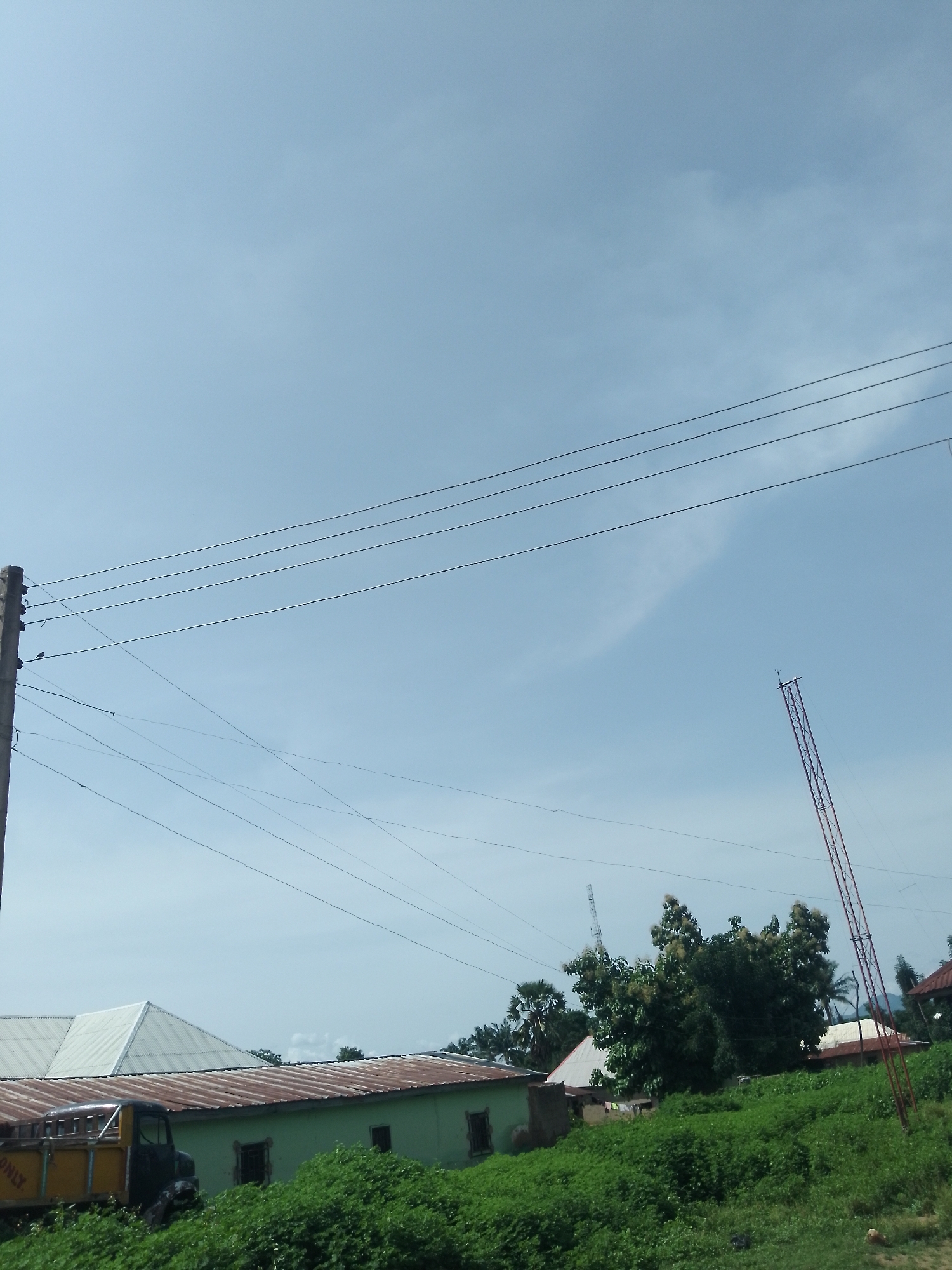The image depicts a scene in Serti, Nigeria, characterized by a clear sky with faint cloud formations. Several power lines are prominently strung across the sky, supported by a concrete utility pole on the left. In the middle ground, there are buildings with corrugated metal roofs, one a light green structure and another with a white, angular roof. A yellow and black truck is parked in front of the green building, partially obscured by dense green vegetation. Further in the background, lush trees and other structures, including a tall, red lattice tower, are visible. The overall environment suggests a semi-urban or rural setting with active electrical infrastructure.