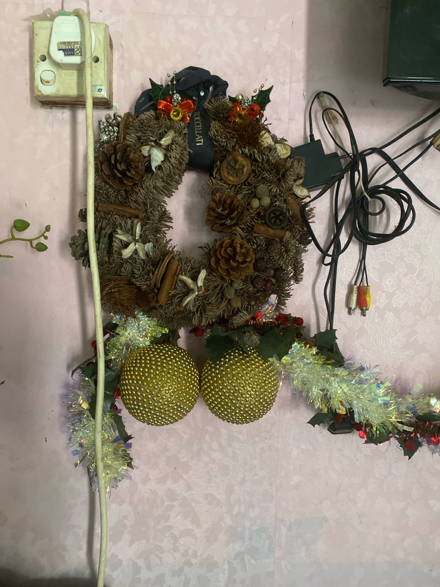 This image depicts a cluttered wall in a room, likely in Batam, Indonesia. A light pink wallpaper with a subtle floral pattern covers the wall. On the left, a white electrical outlet with a white plug and a coiled white cable is mounted on the wall. To the right of the outlet, a circular Christmas wreath made of natural materials like pinecones, cinnamon sticks, and dried fruit hangs on the wall. The wreath is adorned with red berries, holly leaves, a red ribbon with a gold bell, and a dark ribbon with text. Below the wreath, two large, textured golden spherical ornaments are suspended, attached to what appears to be a garland of green tinsel and holly leaves. To the right of the wreath, a bundle of black cables and audio-visual connectors is draped against the wall. The overall scene suggests a residential or office space with festive decorations and practical electrical infrastructure.