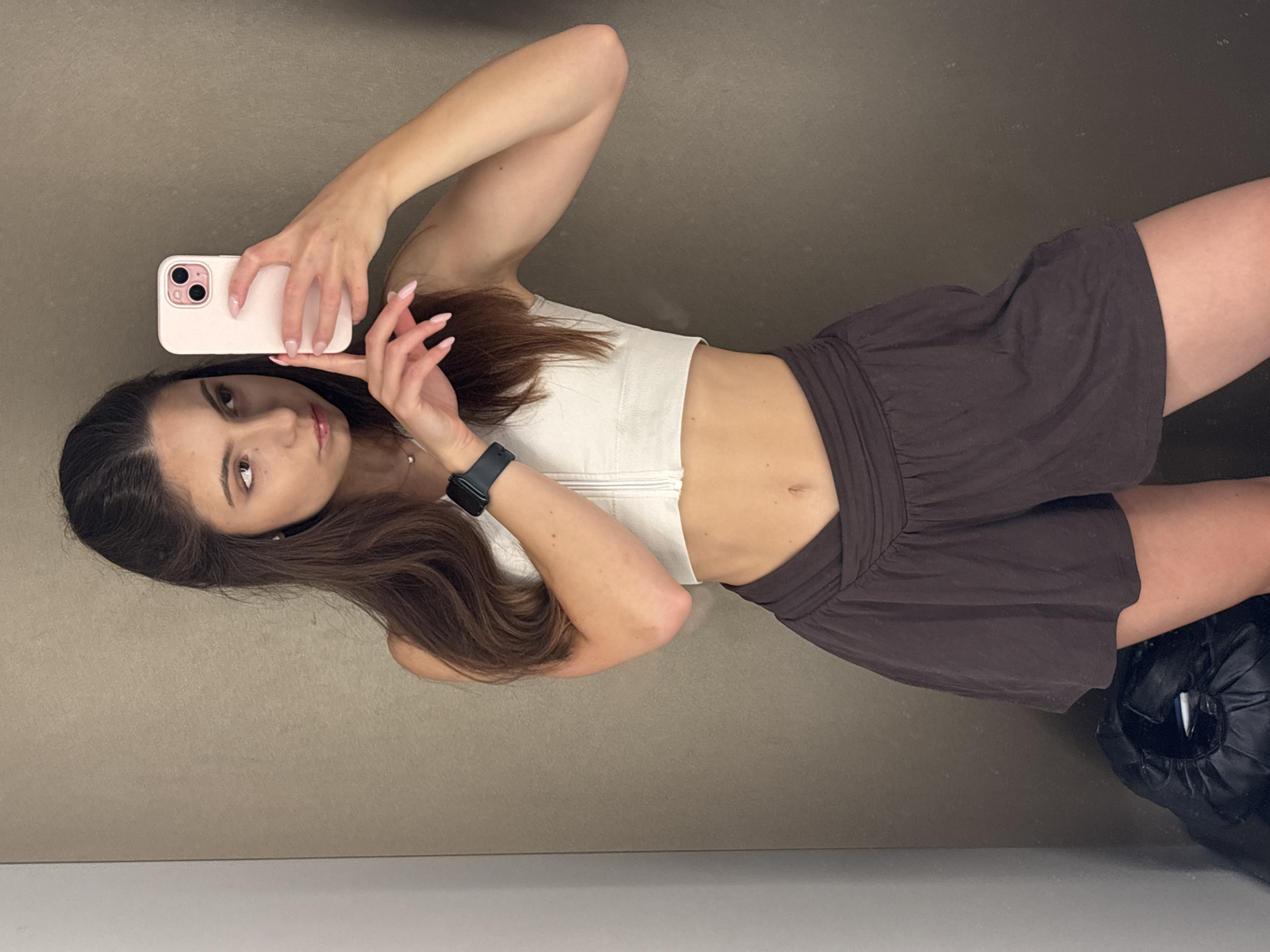 The media file is a selfie taken by a young woman in a fitting room. She is holding a pink smartphone in her left hand, which is angled upwards to capture the reflection. The woman has long, dark brown hair and is wearing a white crop top and dark brown shorts. A smartwatch is visible on her left wrist. The background appears to be the wall of a fitting room, with a gray-brown texture. To the right of the woman, a black garment bag is partially visible. The lighting is even, suggesting an indoor setting. The woman's expression is neutral, and her pose is casual. There is no visible text in the image. The setting of a fitting room, combined with the way the photo is taken, suggests she might be trying on clothes or checking her appearance. The image is oriented vertically, with the woman appearing to be standing.