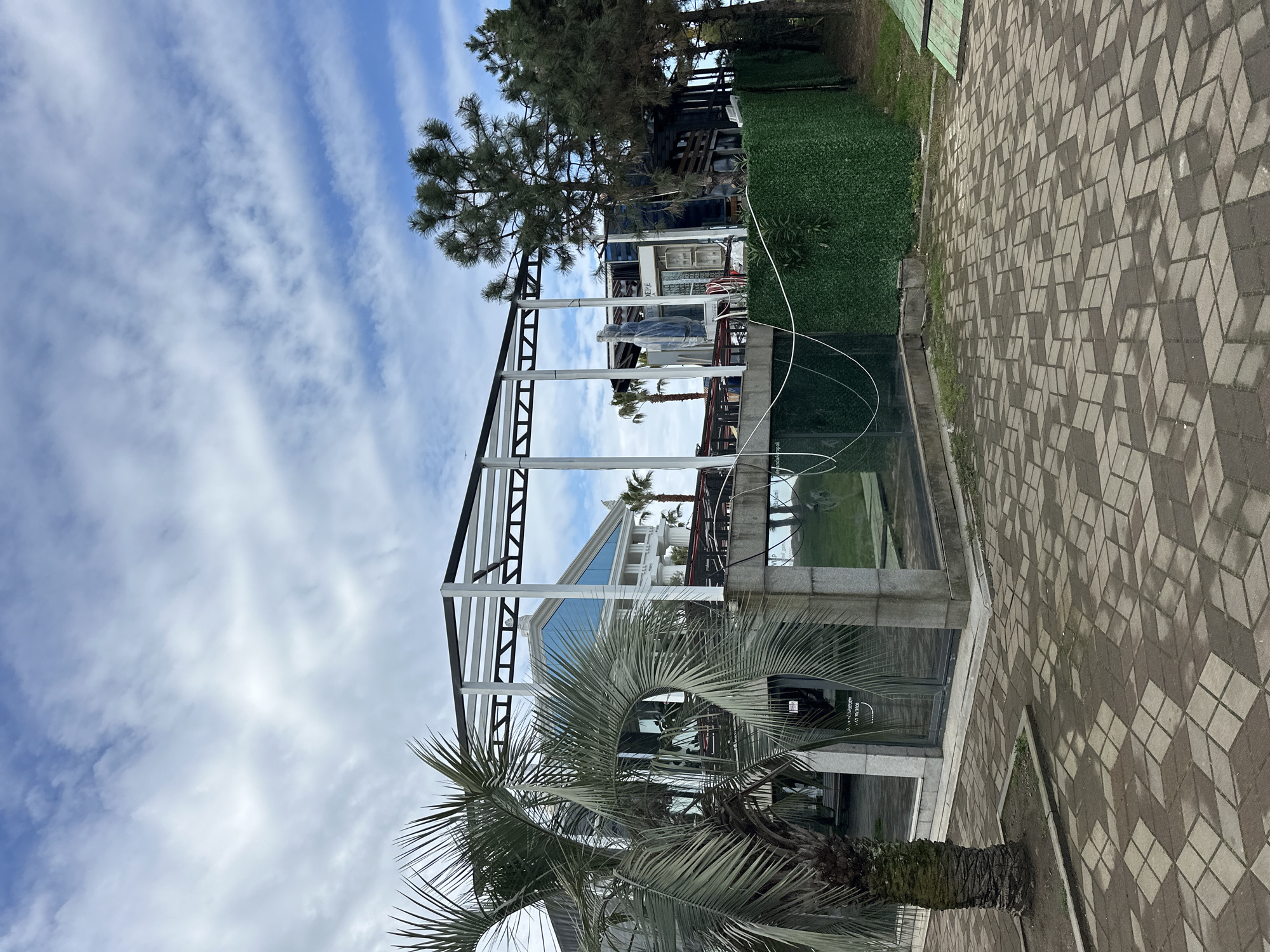 The image shows an outdoor scene in Batumi, Georgia, featuring a partially constructed building with a black and white steel frame. Palm trees are present in the foreground and middle ground. The sky is visible with scattered clouds. A patterned cobblestone walkway is visible on the right side of the frame. Green foliage and a building are visible in the background. The scene appears to be a construction site or an area undergoing renovation.
