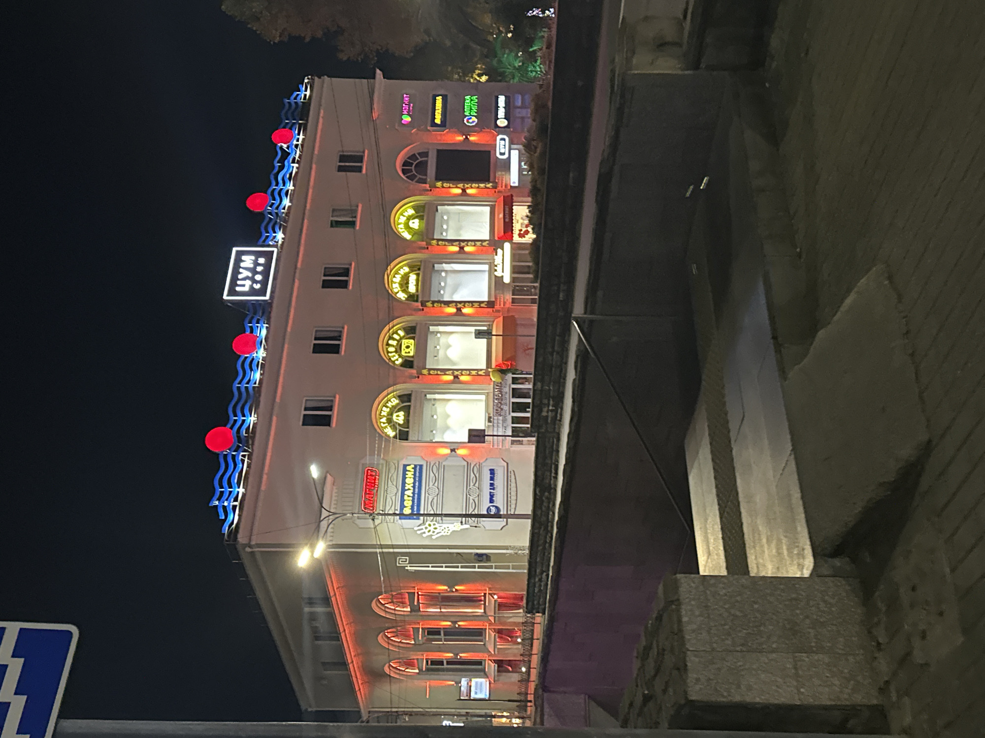 This is a nighttime, eye-level, full shot of a multi-story building in the city of Sochi, Russia, which appears to be a shopping center or department store. The building is illuminated by red and blue neon lights. The most prominent signage is "ЦУМ СОЧИ" (TSUM SOCHI) in large white block letters on a black background, with blue wavy lines on either side and red circles interspersed along the roofline. Smaller signs with Cyrillic text are visible on the facade, indicating various businesses such as "МАГНИТ" (Magnit), "АПТЕКА РИГЛА" (Apteka Rigla - Rigla Pharmacy), and "ОБУВЬ" (Obuv - Shoes). The building has multiple arched windows on the ground floor, many of which are lit from within, revealing empty display areas. The facade is decorated with red accent lighting. In the foreground, a concrete bridge or elevated walkway with stone railings is visible. The area appears to be a public space, possibly a street or square, with paved walkways and some shrubbery. The sky is dark, indicating nighttime.