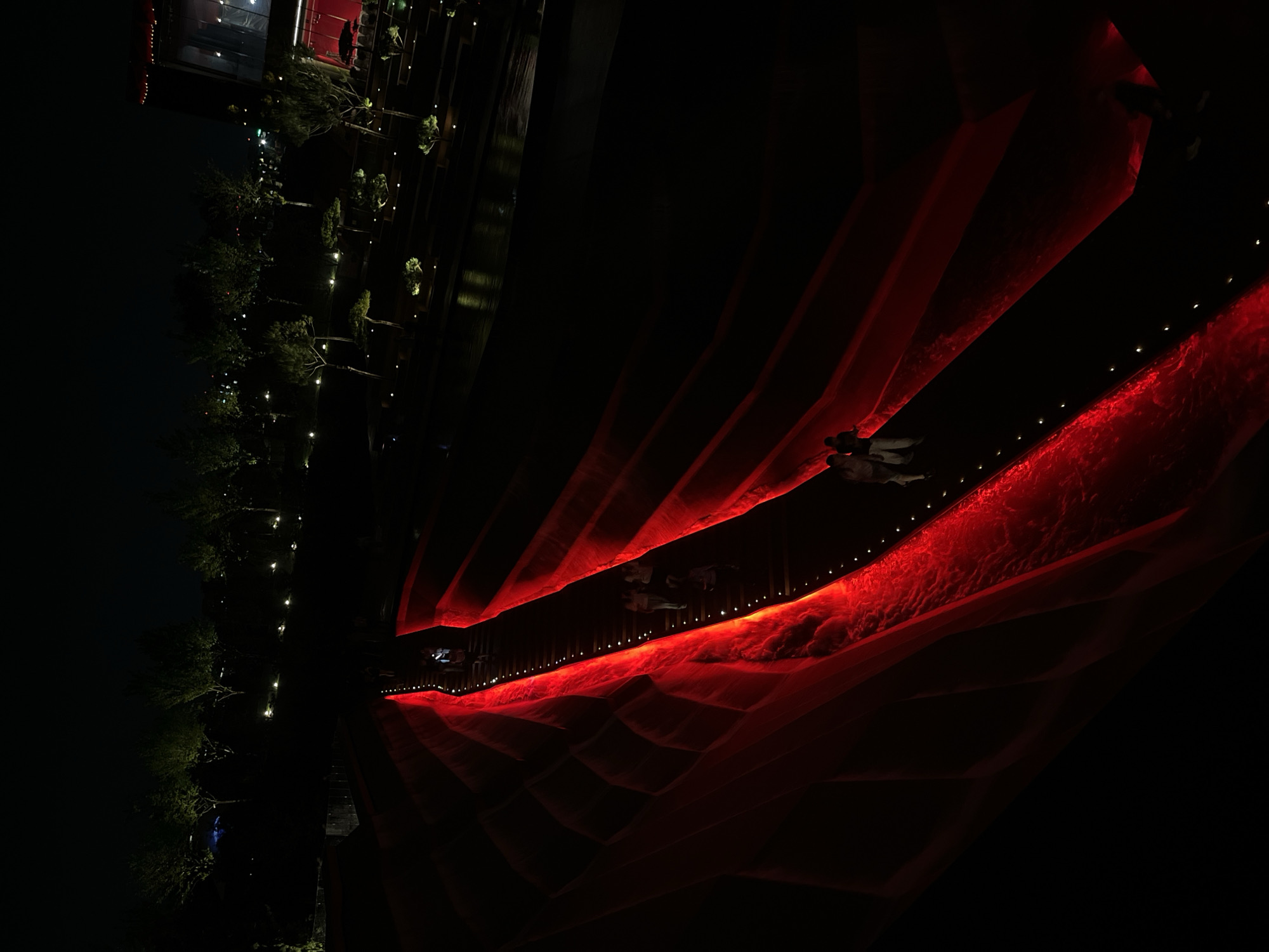This is a nighttime aerial shot of a modern park in Krasnodar, Russia. The park features a striking architectural element: a winding pathway or bridge illuminated by vibrant red light, creating a dramatic visual effect against the dark surroundings. Several individuals are walking along this illuminated path, some in small groups and others alone. The path appears to be elevated and surrounded by dark, possibly water features or landscaping, with scattered small lights visible further in the background. Trees with subtle uplighting are visible in the distance, suggesting a manicured and artistically lit urban green space. The overall impression is one of a contemporary, stylish, and somewhat futuristic public area designed for evening enjoyment.