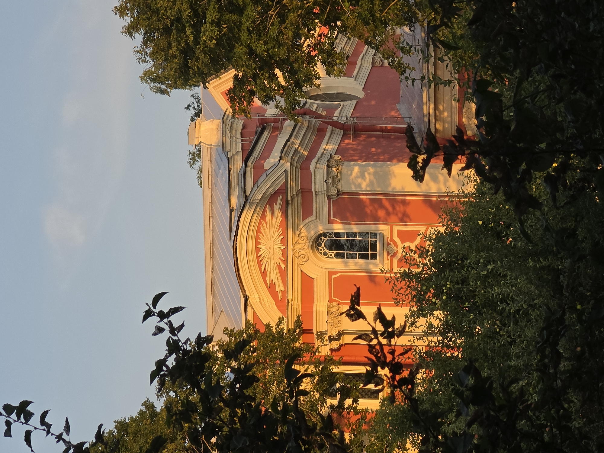 The image shows the facade of a red building with white decorative elements, possibly a church or a historical building. The building is partially obscured by lush green foliage in the foreground and on the sides. The warm, golden light suggests it is either sunrise or sunset, casting long shadows and highlighting the architectural details of the building. The sky above is a pale blue with a few wispy clouds. The building appears to be in Strelna, Russia, judging by the architectural style which is reminiscent of Baroque or Neoclassical design prevalent in the region. The scene is tranquil and serene, emphasizing the beauty of the architecture and its natural surroundings.