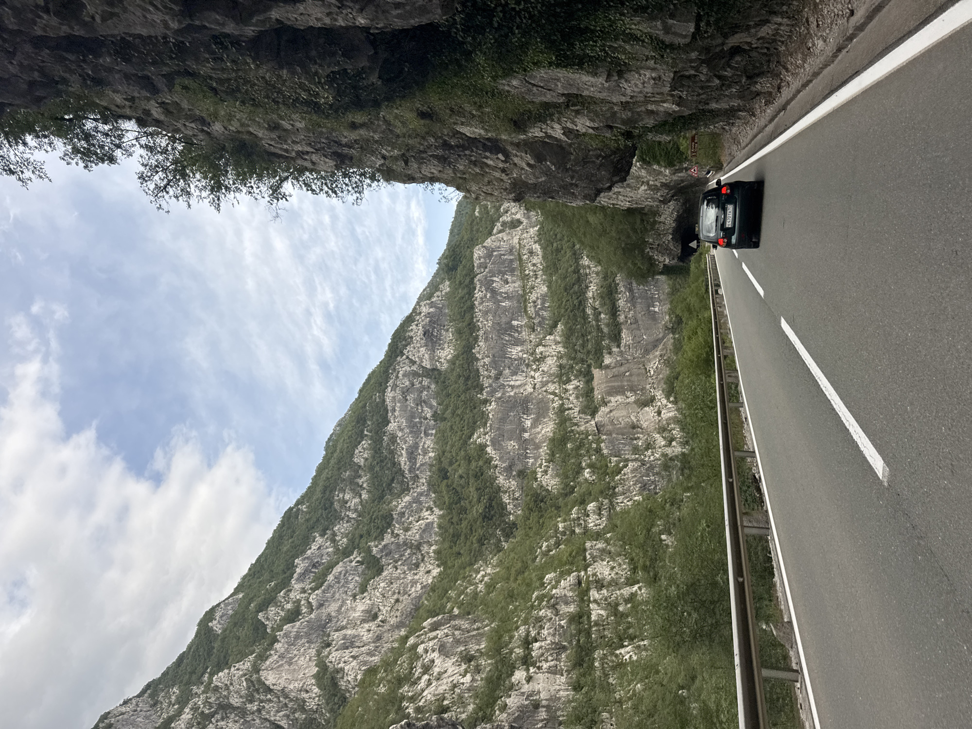 A black hatchback automobile drives along a paved road with white lane markings. The road is bordered by a metal guardrail on the left and a sheer, rocky cliff face covered in vegetation on the right. In the background, lush green mountains rise steeply on either side, with a cloudy sky above. The scene appears to be a scenic mountain pass, possibly located near Vlahovići, Montenegro, a region known for its dramatic natural landscapes. The overall impression is one of travel through a rugged and beautiful natural environment.