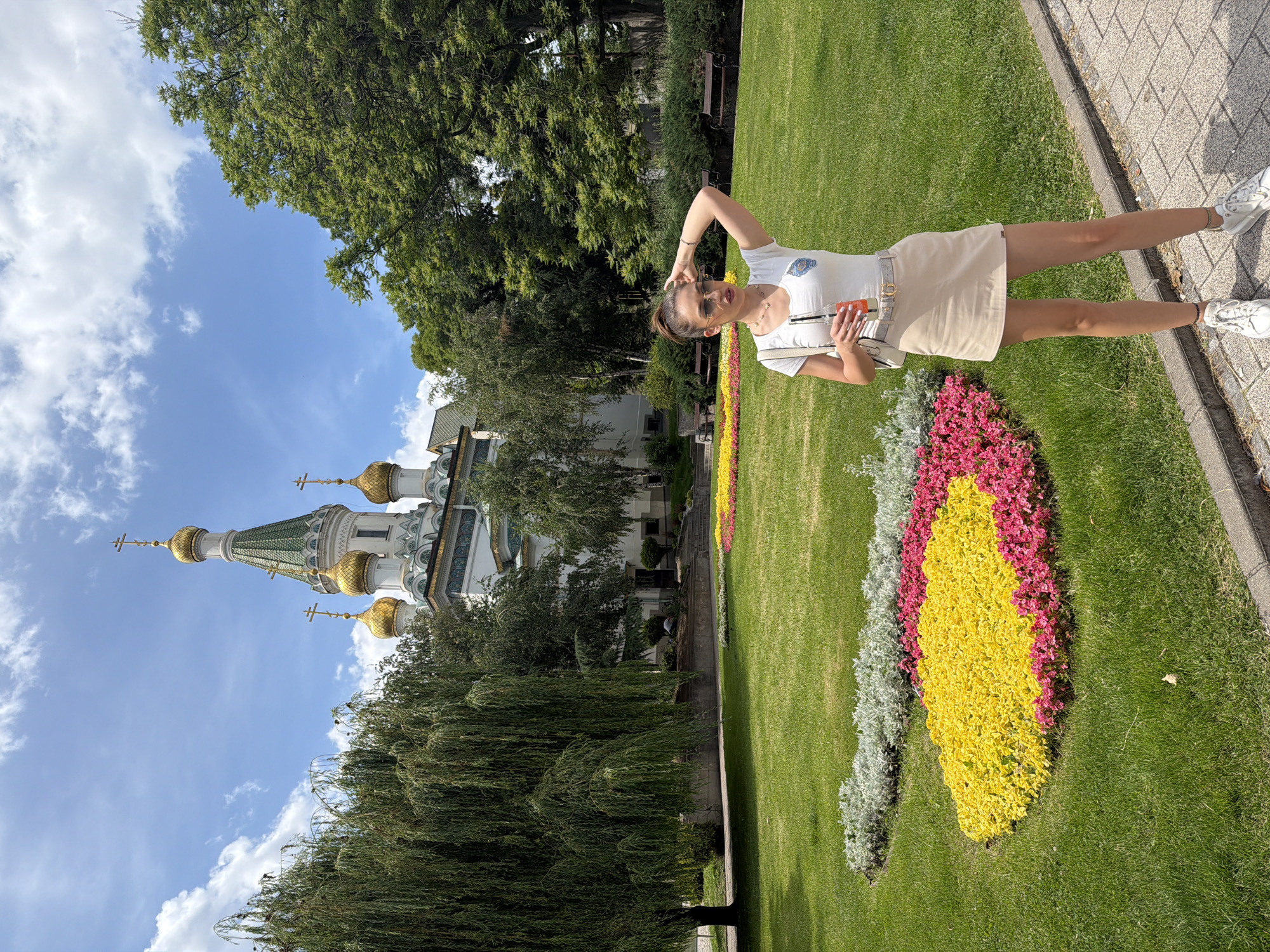 In Sofia, Bulgaria, in front of the Saint Sofia Church, a woman stands on a paved walkway. She is wearing a white t-shirt, a light beige skirt, a beige belt with a gold buckle, and white sneakers. She has sunglasses on and her hair is pulled back in a ponytail. She is holding a bottle with a drink in her right hand and her left hand is on her head. Behind her is a well-maintained park with green grass and flower beds filled with yellow and pink flowers. In the background, the white and gold domes of the church are visible, as are several large green trees and benches. The sky is blue with scattered white clouds.