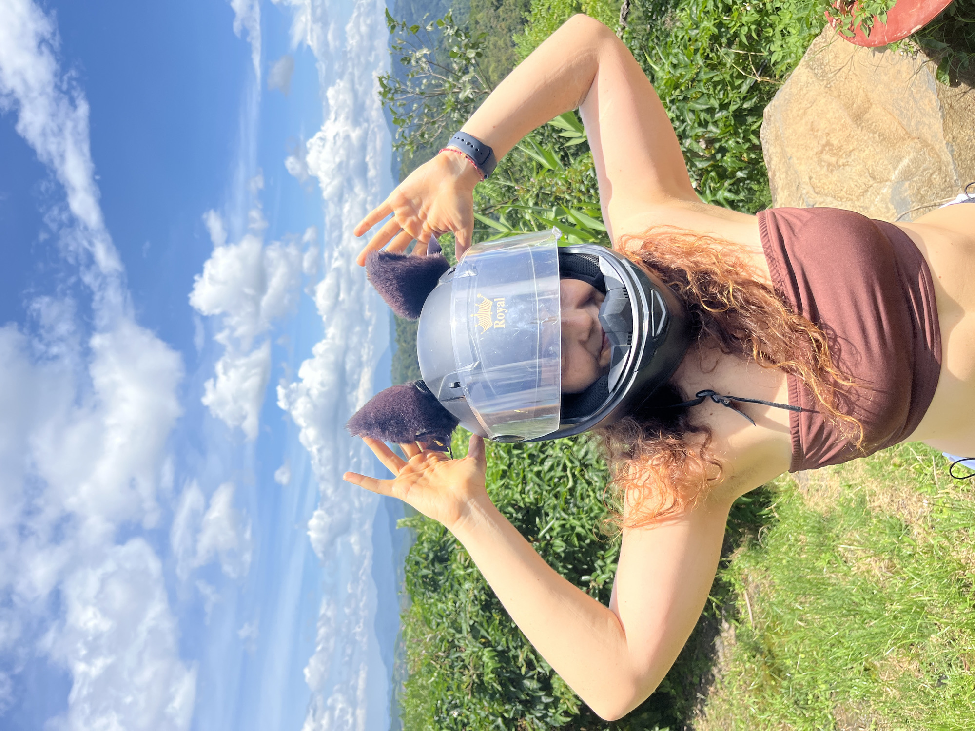A young woman with curly red hair and cat ears attached to a motorcycle helmet smiles at the camera. She is wearing a brown tube top and holding her hands up in a "peace" sign on either side of her helmet. The background is a lush green landscape with mountains in the distance under a bright blue sky with white clouds. The location is in Sơn Điền, Vietnam.