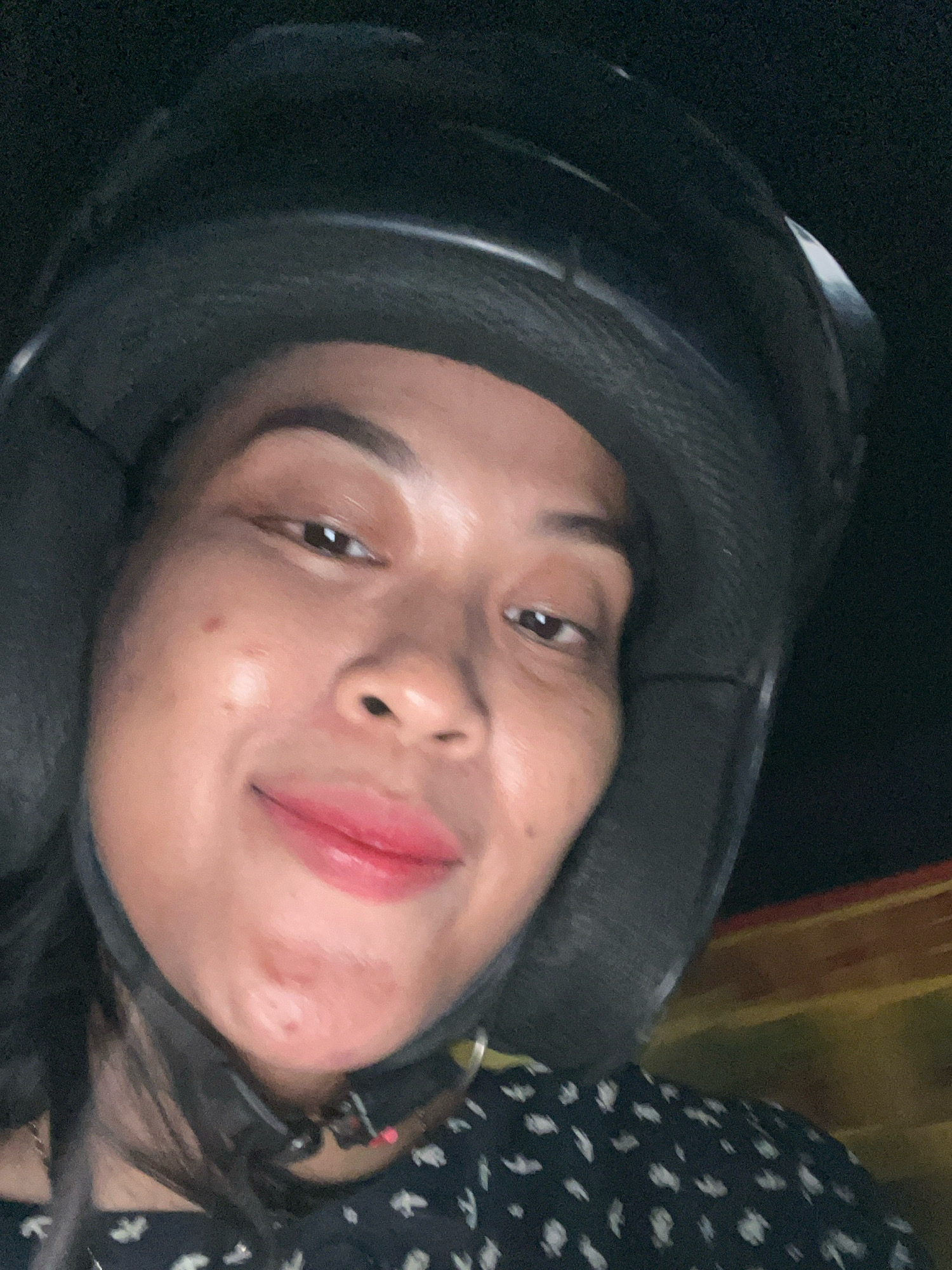 A close-up, selfie-style image shows a woman wearing a black helmet. Her face is the primary focus, with her mouth slightly open in a smile, revealing glossy red lipstick. She has dark hair visible on the sides of her face, and is wearing a dark, patterned shirt. The background is dark, with some blurred yellow and red stripes visible on the right side, suggesting she might be on a moving vehicle in an urban or suburban area of Kecamatan Pati, Indonesia. The lighting appears to be low, possibly from artificial sources or at night.