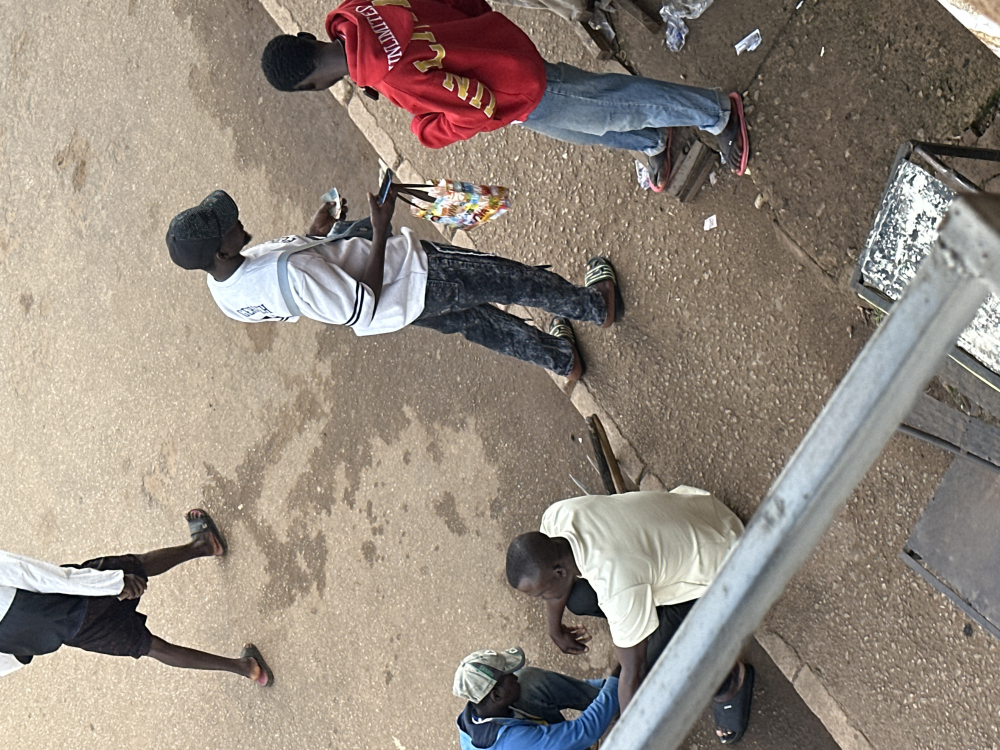 The image depicts a street scene in Bukuru, Nigeria, with several individuals engaged in various activities. In the foreground, a person wearing a black sandal and dark shorts walks away from the camera. To the right, two men stand facing each other. One man, wearing a black cap and a white and grey shirt, holds what appears to be currency in his hands and a colorful bag. The other man is dressed in a red hoodie with yellow lettering that reads "UNLIMITED," and blue jeans. Further to the left, another man in a blue jacket and a green cap is crouched down, possibly interacting with the ground or an object. Beside him, a man in a light-colored t-shirt and dark pants sits on the ground. The environment consists of a paved street with scattered debris, suggesting an outdoor marketplace or a busy thoroughfare. The lighting indicates it is daytime.