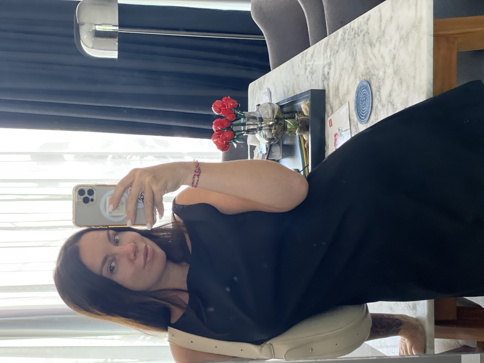 A woman is taking a selfie in what appears to be a hotel room. She is wearing a black dress and a cream-colored shoulder bag. She is holding a smartphone in her right hand, which is angled to capture her reflection. In the background, there is a table with a vase of red flowers, a small succulent plant, and a tray of incense sticks. Dark blue curtains are visible to the right, and sheer white curtains are on the left. The overall impression is that of a clean, modern hotel room. It is not possible to determine the location within Kediri, Indonesia based solely on the visual information provided in the image.