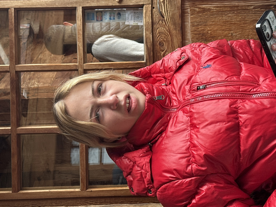 A young individual with blonde hair and light-colored eyes is depicted in a medium-shot, gazing directly into the camera with a slightly open mouth and raised eyebrows. The person is wearing a bright red, shiny puffer jacket with a hood, a prominent front zipper, and a small blue embroidered logo (consistent with a polo rider) on the left chest. A dark-colored smartphone with a patterned case is partially visible in the lower right foreground, held by the individual.
The background consists of a multi-pane wooden window or door frame with distinct wooden mullions and rails. Through the right panes of this window, a reflection is visible showing the back of another individual with a bald or closely-shaved head, wearing a light-colored, possibly ribbed, sleeveless vest or jacket. Below this reflection, various payment card logos (including Mastercard and Visa) and a QR code are discernible, affixed to a surface within the reflected space. The wooden construction and winter clothing suggest a cold-weather environment, consistent with the location of La Clusaz, France.