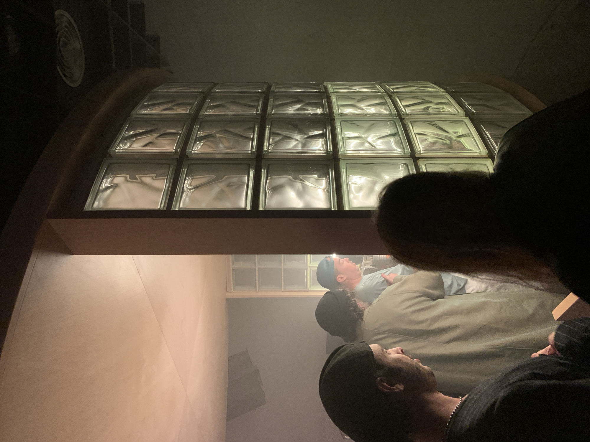 A view through a series of glass blocks shows an interior scene with three people on what appears to be a bed, which reflects the layout of the glass block grid. The angle gives the impression that the viewer is looking from a higher position, possibly through a window or ceiling feature made of the glass blocks. There is one male laying down with a beanie and chain necklace, and two more figures laying behind him. The setting is dark, with the light illuminating the bed area. The floor and walls are pale in color. The location is Tel-Aviv, Israel, as per the query parameters.
