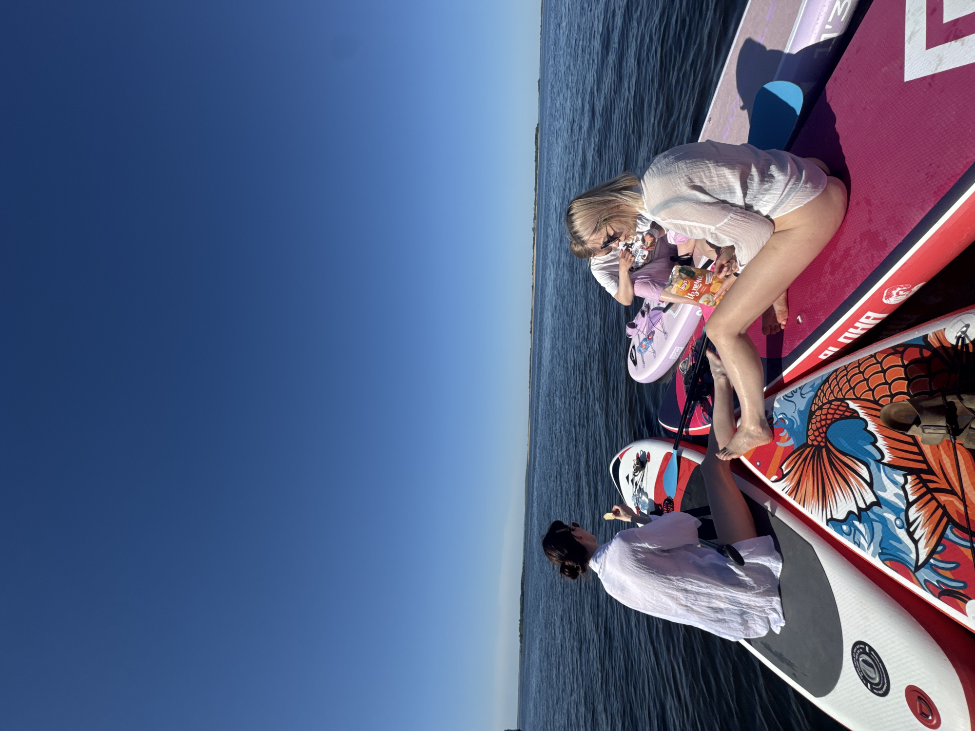 The image shows a group of individuals on paddleboards in a body of water, possibly a lake or river. Two women are visible. One woman is on a pink paddleboard and appears to be snacking from a bag. She is wearing a white, semi-transparent shirt. The second woman is on a separate paddleboard and is also wearing a white shirt. The sky is clear and blue. The presence of the bodies of water and clear sky indicates this could be a warm, sunny day. Given the geographical location, this might be Усть-Курдюм, Russia, as requested.