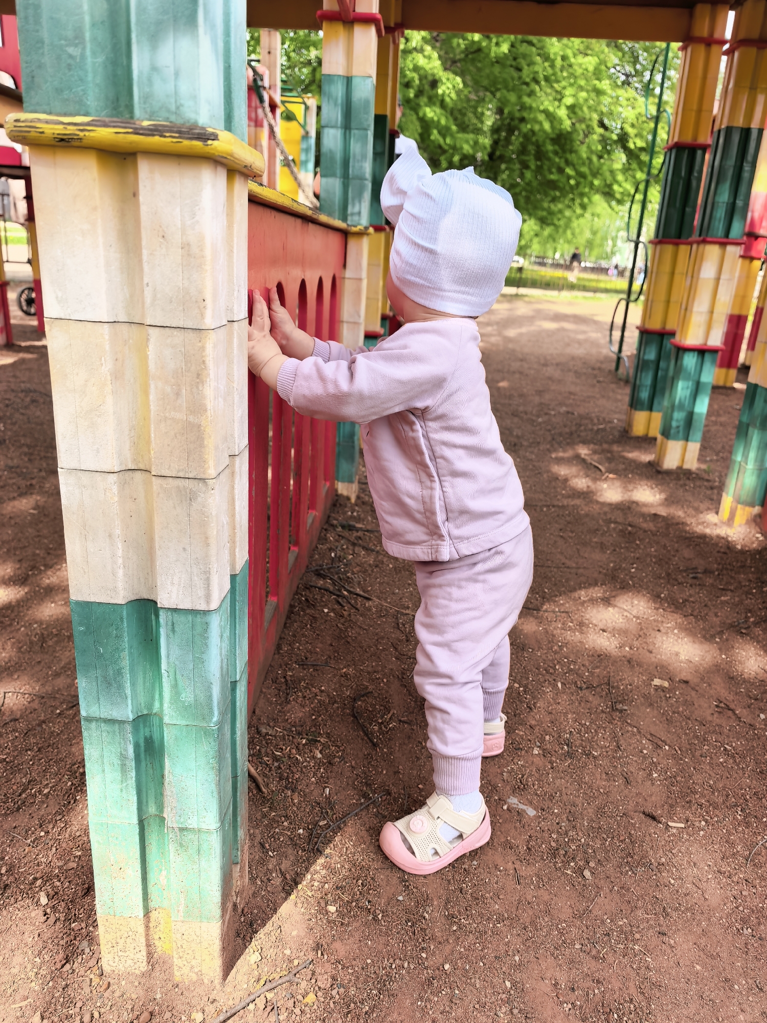 A toddler in a light purple outfit and white cap stands on red-brown dirt at a playground in Sterlitamak, Russia, holding onto a colorful structure. The structure is composed of a yellow-topped, beige-and-green pillar connected to a red fence-like section with arched cutouts. Behind the toddler, more parts of the play structure are visible, including yellow and green sections and other colorful pillars. The playground is located in a park with green trees in the background.