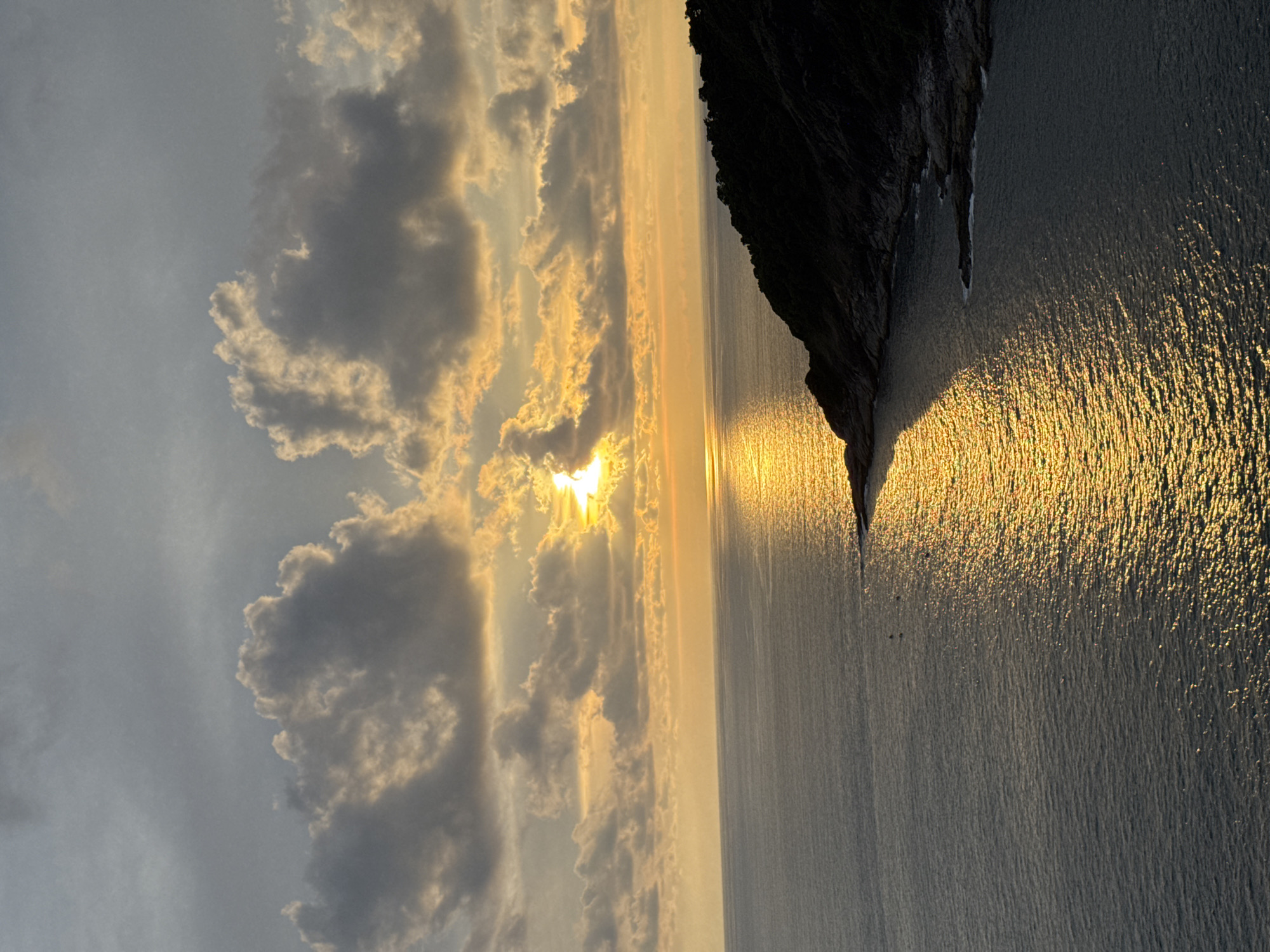 The image shows a sunset over the ocean at Rawai, Thailand. The sun is partially obscured by clouds, casting a golden reflection on the water. A dark, tree-covered headland juts out into the sea on the right side of the frame. The sky is a mix of grey and orange clouds. There are no people or specific objects of interest visible in the scene, only the natural landscape.
