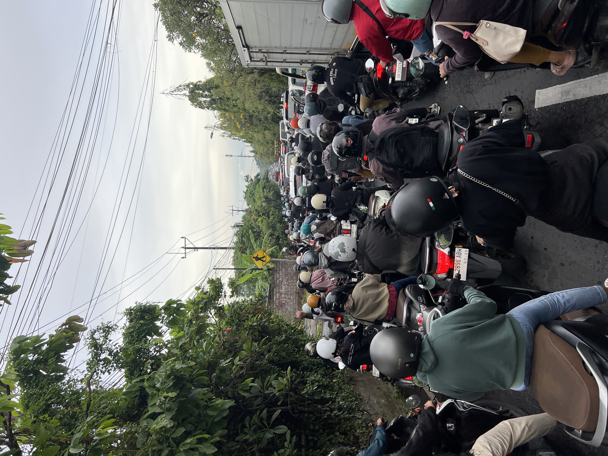 A congested road in Denpasar, Indonesia, shows a dense traffic jam of motorcycles, with riders wearing helmets. The scene is filled with motorcycles closely packed together, a mixture of light-colored and dark-colored helmets on riders, and vehicles, including a box truck, barely visible in the background. Overhead, a network of power lines is visible against a cloudy sky. Lush green foliage lines the roadside.