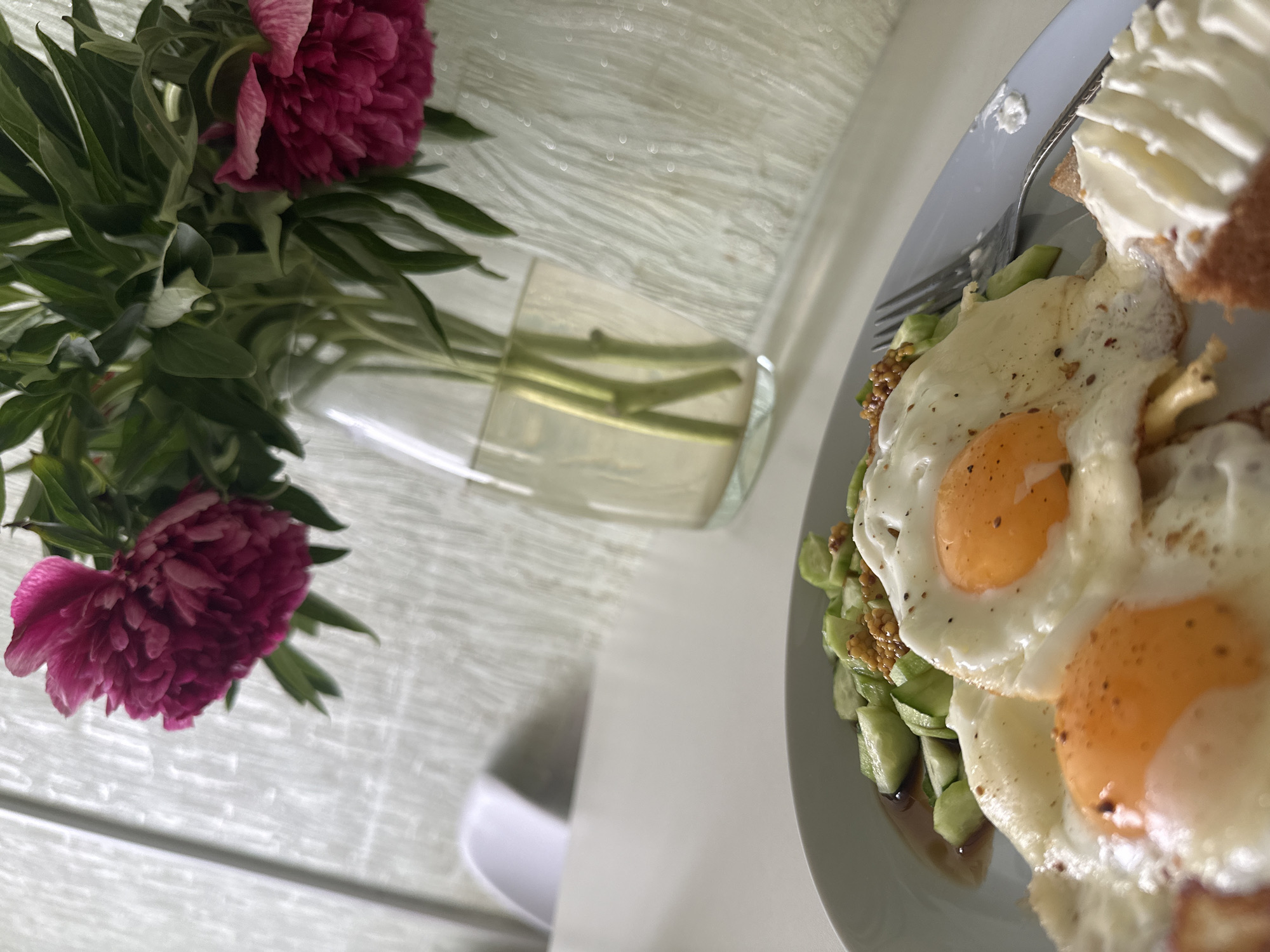The image captures an angled, close-up view of a tranquil tabletop scene, likely an indoor setting in Chaikovsky, Russia.
In the foreground on the right, a white plate holds a meal featuring two perfectly fried eggs with bright orange yolks and crisp whites. Beneath the eggs, there's a bed of chopped green vegetables, possibly cucumber or avocado, dressed with a darker sauce and sprinkled with small, yellowish seeds, resembling mustard seeds or nuts. To the upper right of the eggs, a piece of toast or bread is topped with a generous, swirling dollop of white cream, likely cream cheese or whipped butter. A silver fork is partially visible on the plate, resting near the food.
To the left, a vibrant bouquet of deep magenta or pink peonies adds a splash of color. The lush flowers, with their many petals, are arranged in a clear glass vase, allowing their green stems to be seen through the water. The surface beneath the vase is a light-colored table, possibly white or light gray, with a subtle textured pattern visible under the vase. A blurred white object, possibly a cup or bowl, is in the lower-left background.
The soft, natural lighting suggests it is daytime, possibly morning or early afternoon, contributing to the serene atmosphere. The scene evokes a sense of comfort and freshness, depicting a pleasant breakfast or brunch. There is no visible text in the image.