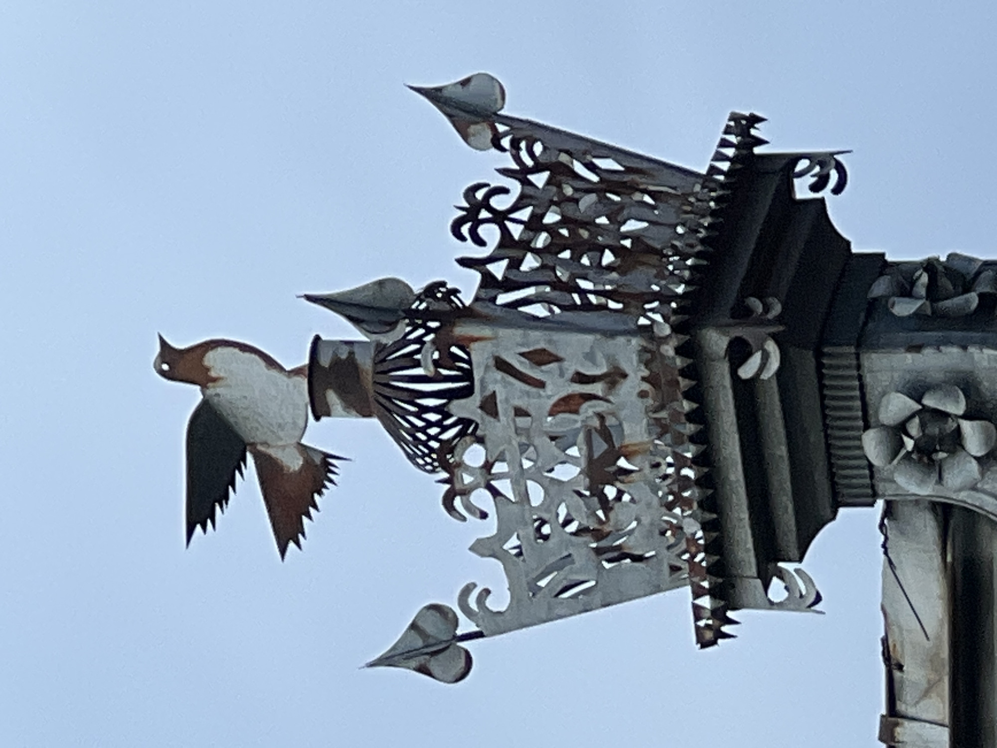 This close-up, angled shot captures a decorative metal sculpture against a pale, overcast sky. The sculpture appears to be a weather vane or part of an architectural embellishment. The main subject is a stylized metal bird, predominantly white with brown markings on its head, wings, and tail. Its wings are spread wide, with sharp, pointed feather details. The bird is attached to a more elaborate section of metalwork featuring intricate cutouts with floral and geometric patterns. This section also includes a jagged, saw-like edge and pointed finial elements. Further to the right, the sculpture transitions into darker, more solid metal components, including what looks like a decorative flower or rosette and ribbed vertical sections. The overall impression is one of weathered metal, suggesting it has been exposed to the elements. The lighting is soft, indicating it's likely daytime, possibly a cloudy or hazy afternoon. There are no visible people or text. The location context is Krasnodar, Russia.