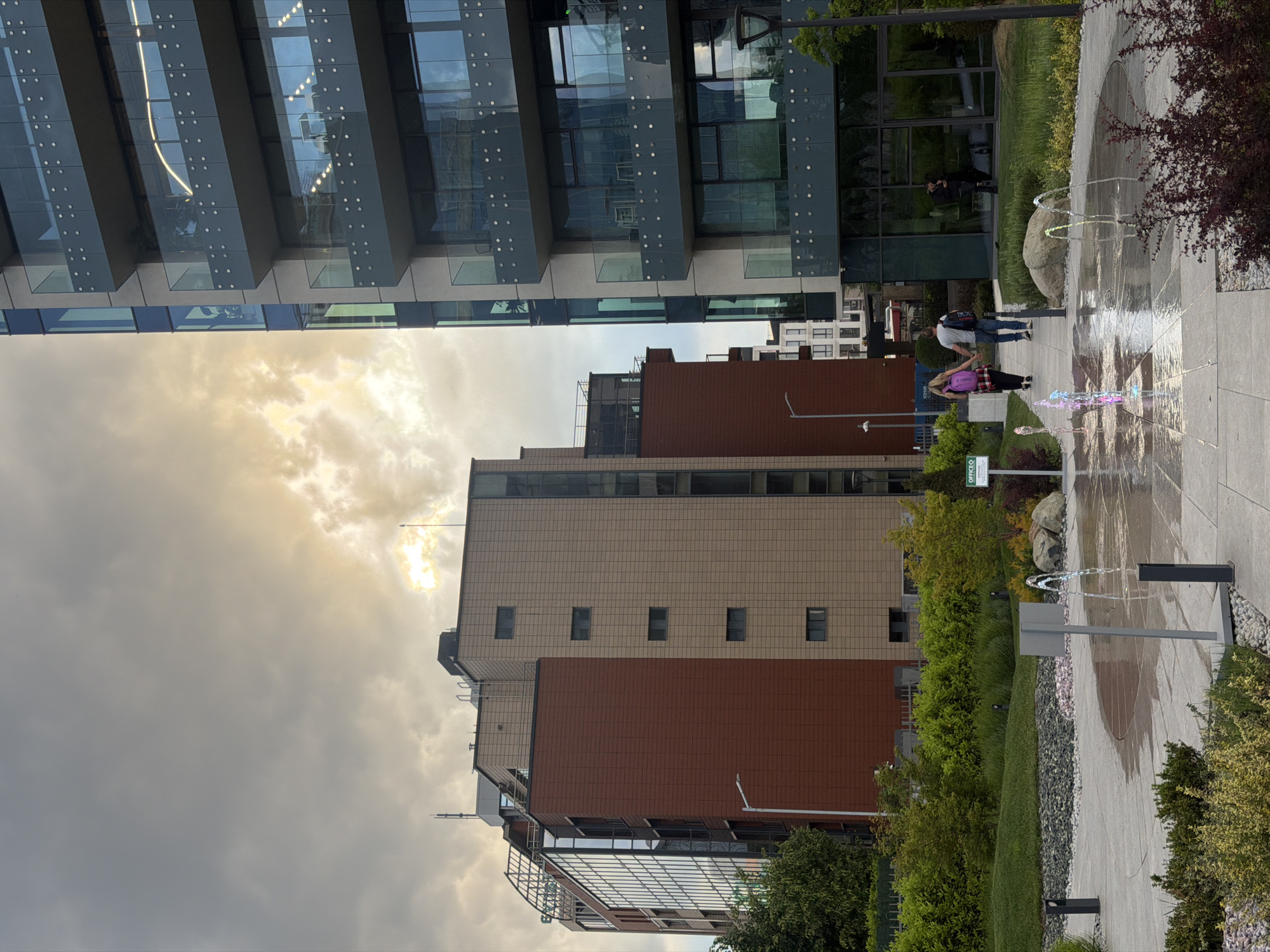 The image captures an urban landscape in Sofia, Bulgaria, featuring modern architecture and a public recreational area under a partially cloudy, sunlit sky.
Dominating the left side of the scene is a tall, contemporary building with multiple stories of glass facades and prominent horizontal structural elements. Its reflective surfaces mirror parts of the sky and the surrounding environment. Below this building, a person is visible reflected in the glass.
In the mid-ground, a multi-tiered building with distinct sections of brown and beige tiling stands, characterized by small, evenly spaced windows. Adjacent to this building, on the right, is a paved public space with an active ground-level fountain. Multiple jets of water spray upwards, and some of these jets are illuminated with colorful lights, displaying hues of pink and purple. Two people are walking on the paved pathway next to the fountain: one individual in a light shirt and dark pants with a backpack, and another wearing a dark top, a plaid skirt, and a vibrant pink backpack.
The area is interspersed with green elements, including manicured lawns, bushes, and trees, creating a park-like atmosphere. The sky above is mostly overcast, but bright sunlight pierces through the clouds in several areas, particularly on the upper left and right, suggesting either a late afternoon or early evening time.
A small green signpost with white lettering is visible near the central building, clearly displaying the text "OFFICEC". The overall impression is of a modern commercial or office district integrated with attractive public amenities.