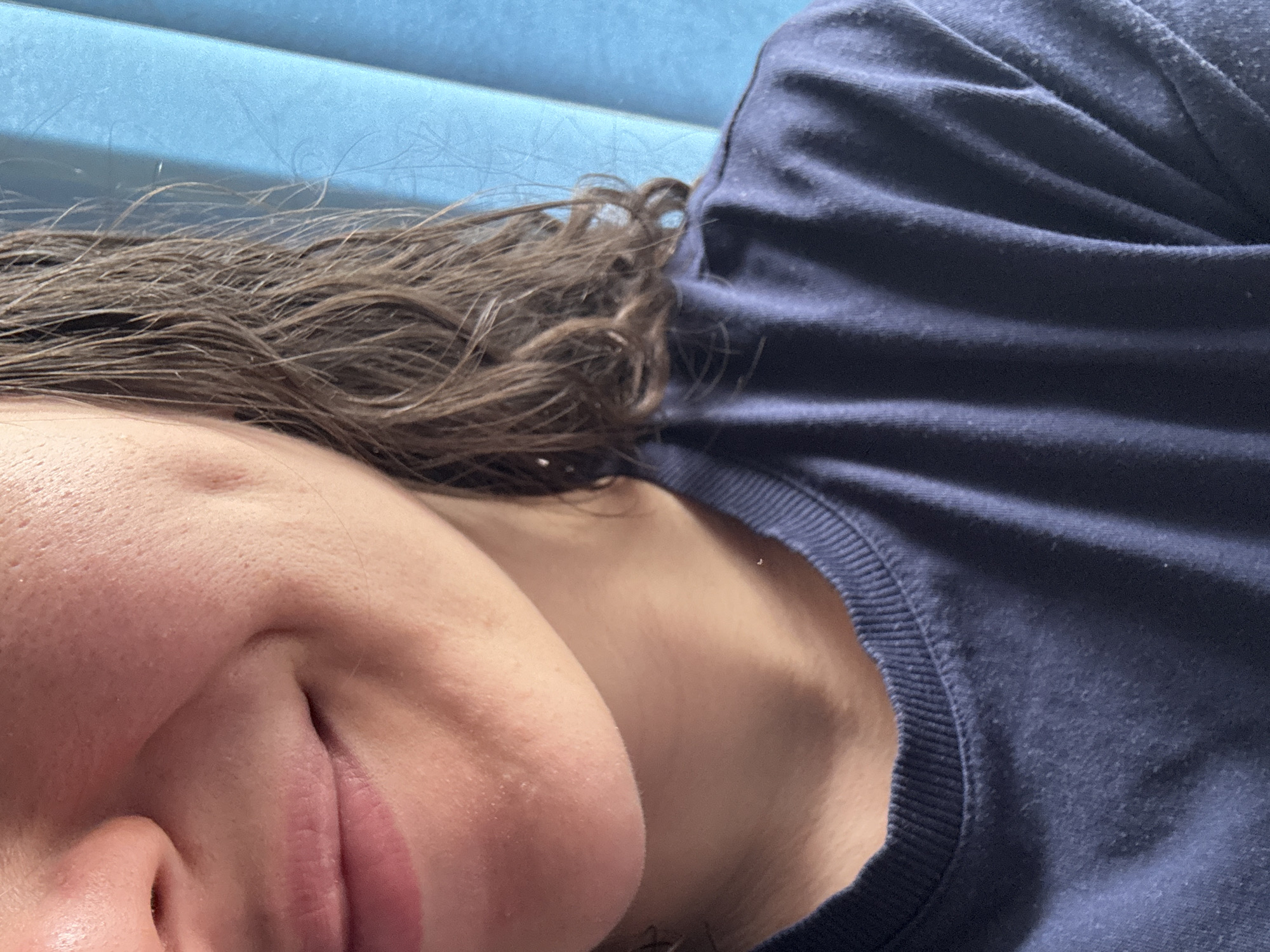 A close-up, indoor shot shows a person lying down, seen from an angled perspective. The person has brown, slightly damp hair. A portion of their face is visible, including the nose, mouth, and cheek. They are wearing a dark blue shirt. The background consists of a blurry, light blue surface.
