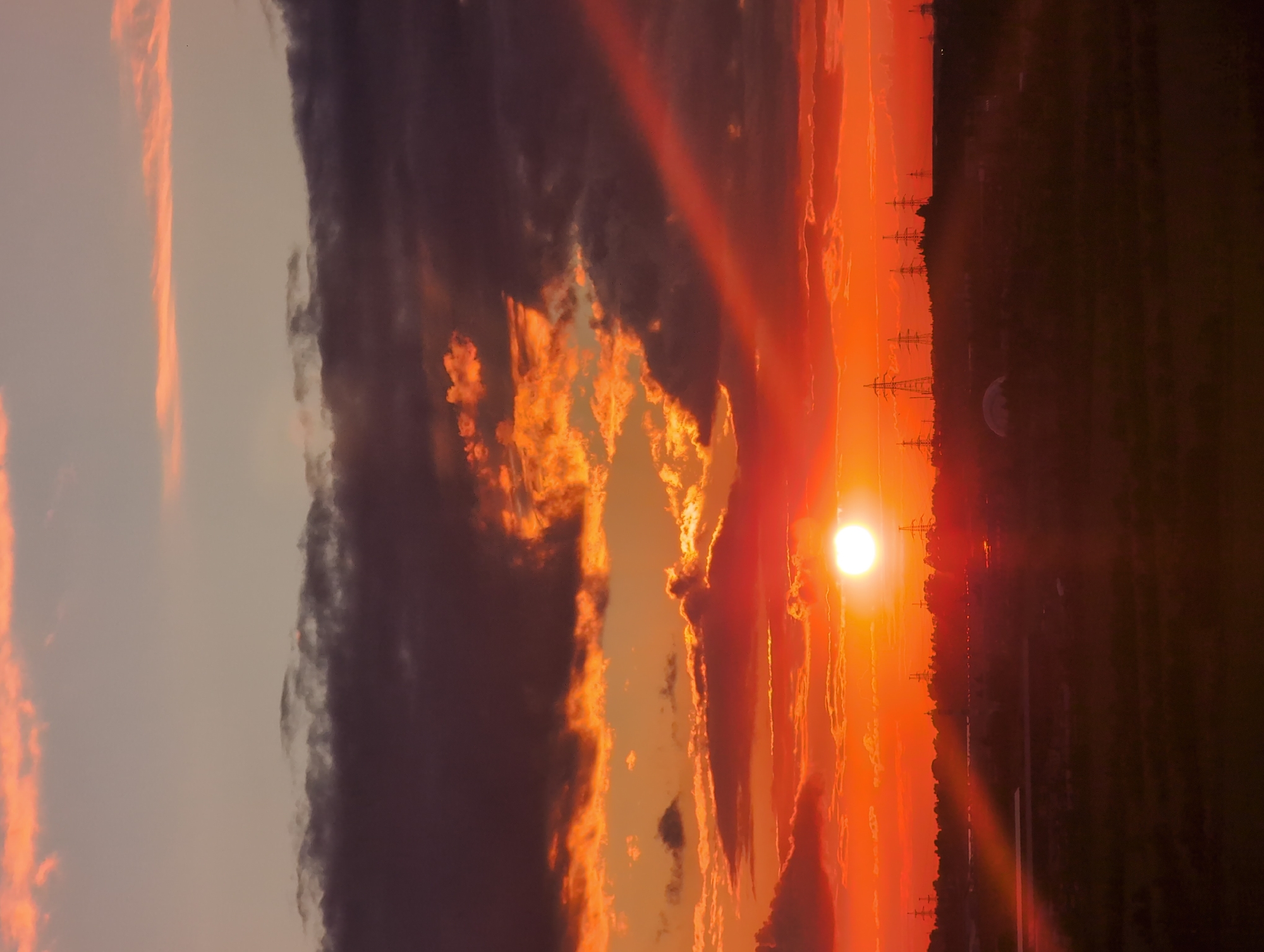 This media file depicts a sunset over a rural landscape in Bugry, Russia. The sun, a bright orb of light, is setting on the right side of the frame, casting a vibrant orange and red glow across the sky. Wispy clouds, tinged with fiery hues, are scattered throughout the sky, creating a dramatic and colorful scene.
In the foreground, silhouetted against the sunset, are several tall power line pylons, suggesting the proximity of electrical infrastructure. Beyond the pylons, a dark, tree-lined horizon stretches across the landscape, with a faint hint of distant lights that could indicate a settlement or further infrastructure. The overall atmosphere is one of natural beauty and tranquility, with the intense colors of the sunset evoking a sense of awe. The image appears to be a vertical photograph, captured during the late evening hours as the sun descends below the horizon.