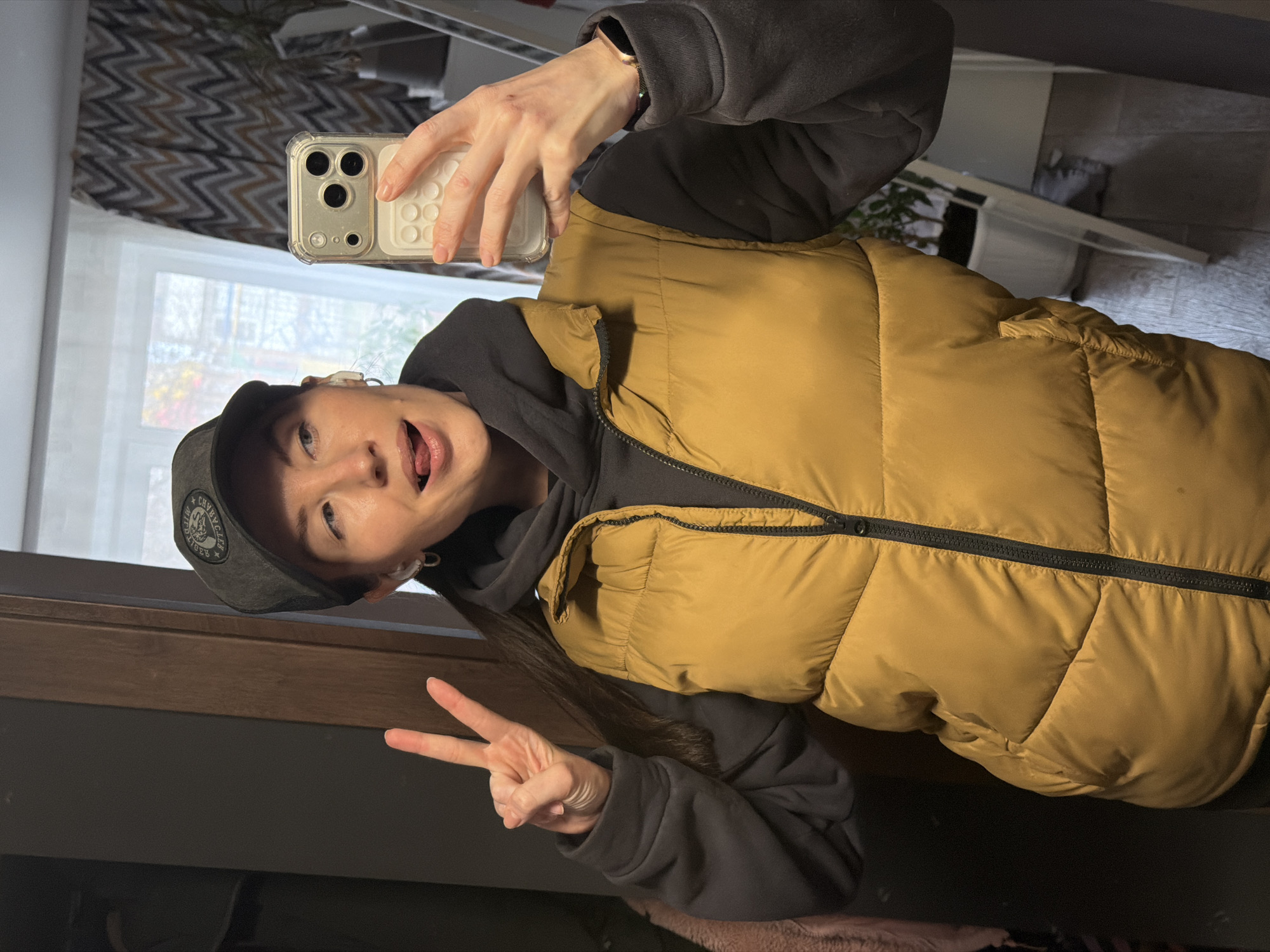 A playful mirror selfie captures a young woman indoors. She has long, dark hair, blue eyes, and is wearing a dark grey baseball cap, a dark grey hooded sweatshirt, and a mustard yellow puffer vest. On her right wrist, she wears a smart watch. She holds a smartphone with a clear case in her right hand to take the photo, while her left hand is raised in a peace sign. Her expression is lighthearted, with her tongue slightly out.
The cap features a black patch with white text: "CHRYSLER" is prominent, with "TIGER" visible below it, and additional text around the edge. The phone case has a textured white area with circular indentations.
The background, reflected in the mirror, shows a window letting in bright daylight, indicating the photo was taken during the day. A curtain with a grey and white zigzag pattern hangs above the window. Other reflections hint at an indoor setting with furniture. The implied location context for this casual moment is Perm, Russia.