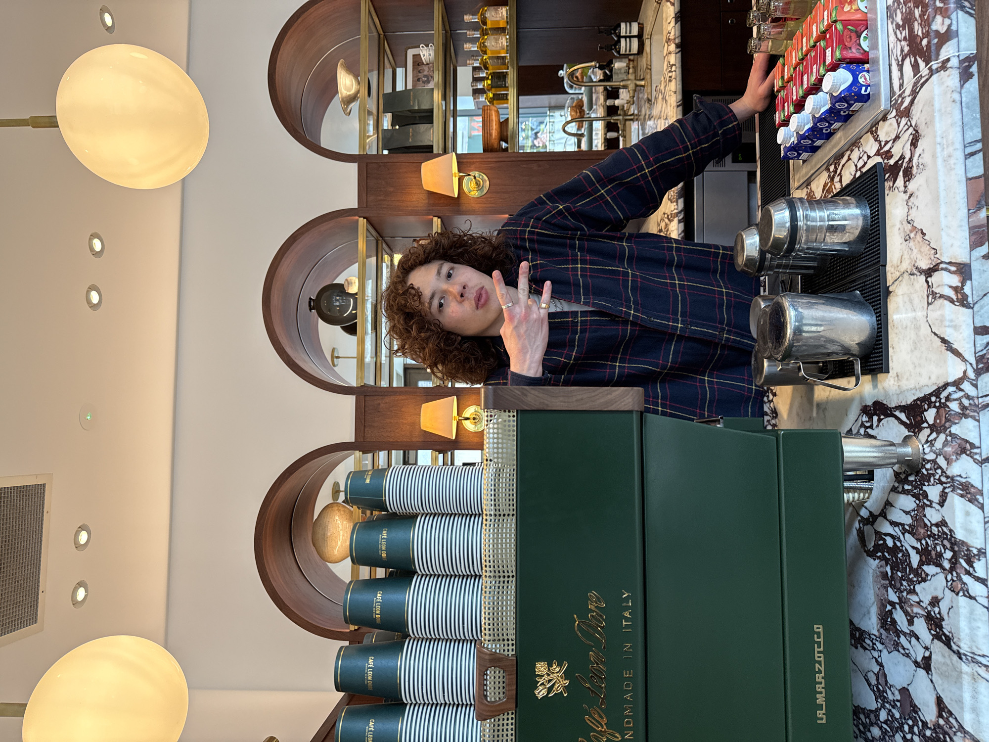 A person with curly brown hair, wearing a blue, red, and yellow plaid shirt, is captured posing playfully at a bar in what appears to be an upscale cafe in London. The individual looks directly at the camera, making a peace sign with their left hand, which is adorned with rings. Their right hand is extended towards a display of juice boxes, some red and others blue and white with a visible "U" logo, alongside small milk cartons.
The setting features a prominent counter made of white and reddish-brown veined marble. Behind the person, dark wood shelving with curved compartments holds various bottles and decorative wall sconces, creating an elegant backdrop. The ceiling is white, fitted with recessed lights and a large, spherical pendant light emitting a warm glow.
In the foreground, a dark green espresso machine is clearly visible, featuring gold lettering that reads "Café Leon D'Or" and "HANDMADE IN ITALY." The brand "La Marzocco" is also discernible on the machine. Stacks of paper cups, also branded "CAFÉ LEON D'OR," are neatly arranged on top of the machine, indicating its use in a commercial setting. The interior is well-lit, suggesting a vibrant atmosphere during what appears to be a daytime operation.