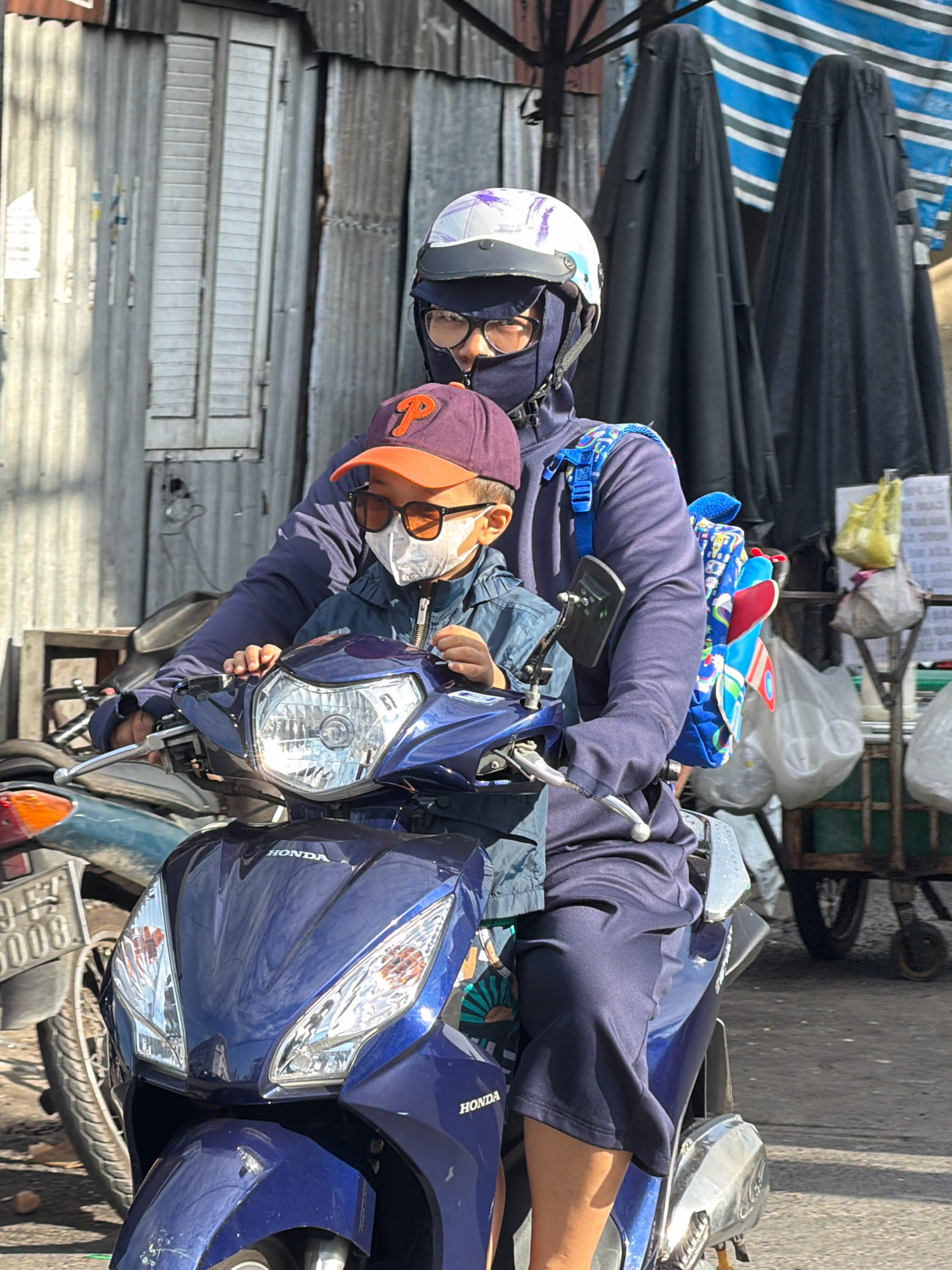 In Nha Trang, Vietnam, a person wearing a blue jacket, dark face mask, and a white and purple helmet is riding a blue scooter, carrying a child in front. The child is wearing an orange-brimmed baseball cap, sunglasses, and a white face mask. The child is also wearing a denim jacket. A colorful backpack is strapped to the person's back. The scooter has the "HONDA" logo visible on its front. In the background, there is a corrugated metal wall and two large, dark, tent-like structures draped with blue and white striped fabric. A market stall with bags and yellow items is also visible on the right side of the image. The ground appears to be paved.