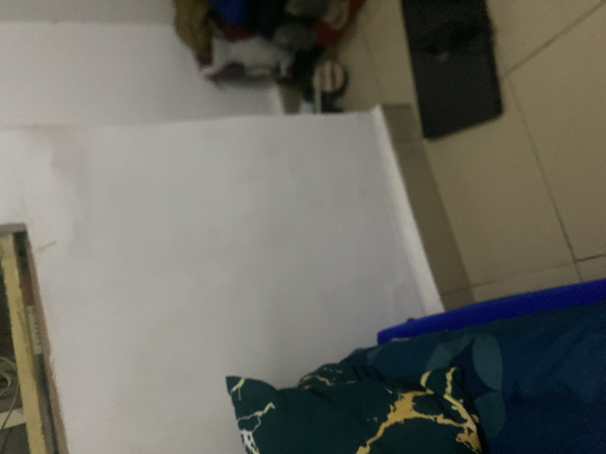 The image depicts an indoor scene, possibly a bedroom or living area. In the foreground, a dark blue pillow with a green and yellow marbled pattern rests on a blue surface, partially obscuring the lower left corner. The wall behind the pillow is a plain, light color, with a beige baseboard running along the bottom. To the right of the wall, a cluttered area shows various items, including what appears to be clothing and footwear. A dark rectangular object, possibly a mat or a thin box, is placed on the tiled floor in the lower right quadrant. The lighting is dim, and the overall image has a grainy quality. The location is not specified in the image.