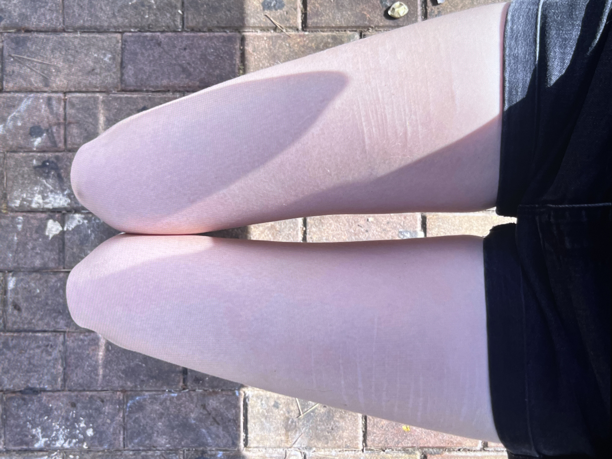 The image shows a close-up view of a person's legs, which are covered in sheer, pale pink tights. The legs are resting on a brick pathway, which appears to be outdoors. Sunlight casts a shadow across the left leg, creating a gradient of light and shade. The person is wearing black shorts that come up to the mid-thigh. The scene is likely set in Torrevieja, Spain, as indicated by the location context, suggesting a warm, sunny day. The overall impression is casual and relaxed. There is no visible text in the image.