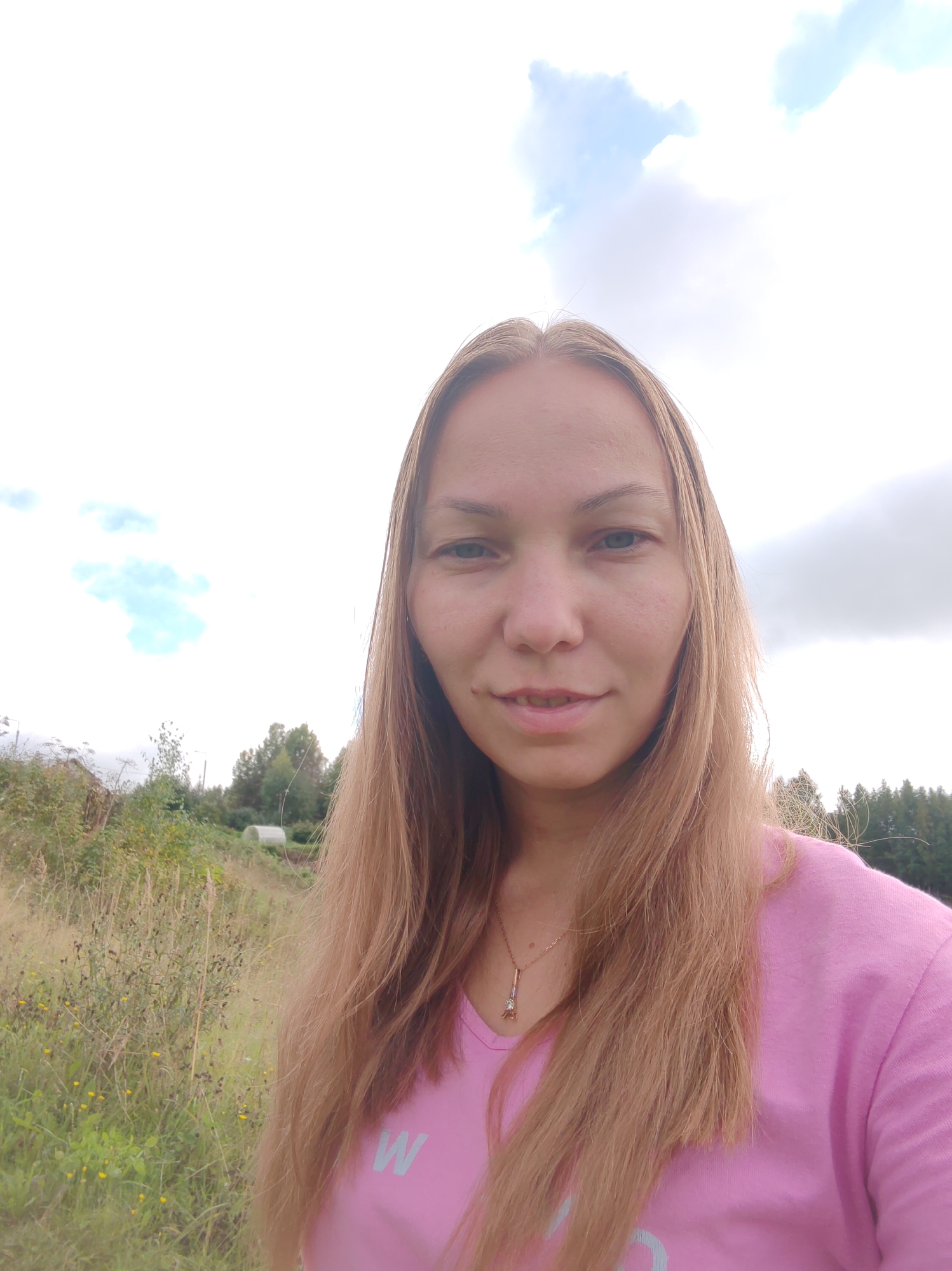 A fair-skinned woman with shoulder-length blonde hair stands in a field of dry grass and wildflowers. She is wearing a bright pink t-shirt with a light blue "W" logo on it, and a delicate gold necklace with a small pendant. Her expression is neutral, with a slight smile. In the background, there are trees, a small building that appears to be a greenhouse, and a cloudy sky. The location is identified as Chukhlem, Russia.
