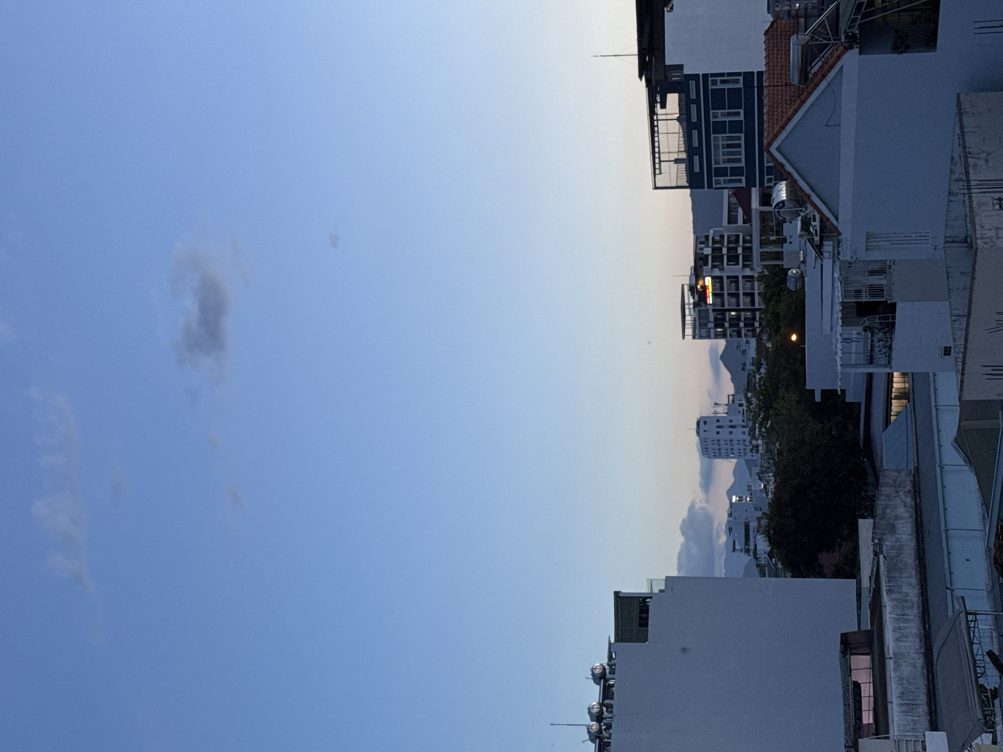 The image depicts a skyline in Bac Nha Trang Ward, Vietnam, at dusk or dawn. Buildings of varying heights and designs line the right side of the frame, ranging from multi-story structures to smaller houses. The sky occupies the majority of the image, transitioning from a deep blue at the bottom to a lighter shade at the top, suggesting the fading light of either sunset or sunrise. A few scattered clouds are visible against the blue sky.