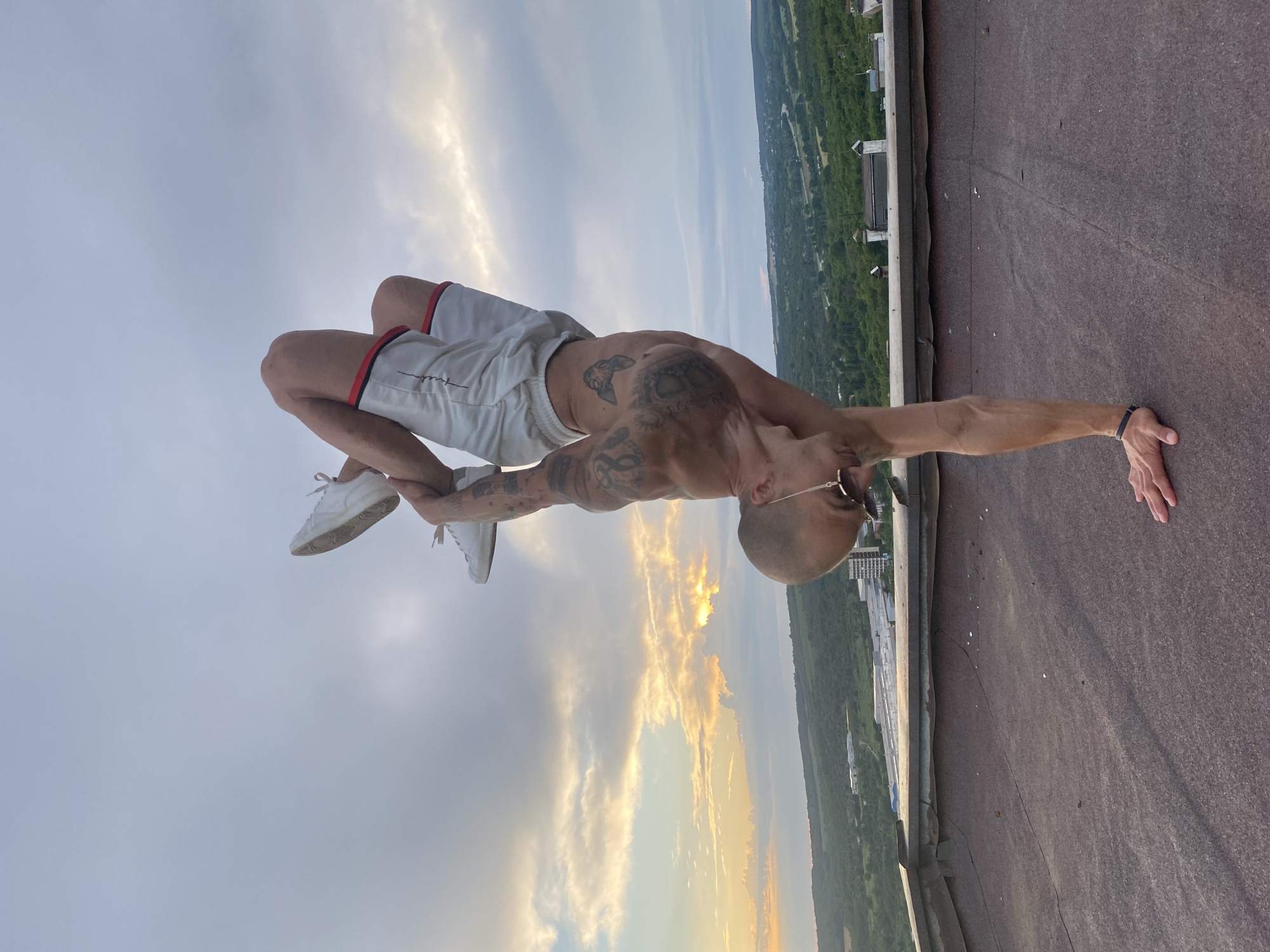 A tattooed, bald man is performing a handstand on a rooftop in Pleven, Bulgaria. He is wearing white shorts with red trim and white sneakers. The man's body is positioned vertically, with his hands on the rooftop and his legs extended upwards, bent at the knees. The background shows a cloudy sky with hints of sunset light, and a green, hilly landscape with buildings in the distance, consistent with the outskirts of Pleven. The rooftop is covered with a dark, textured material.