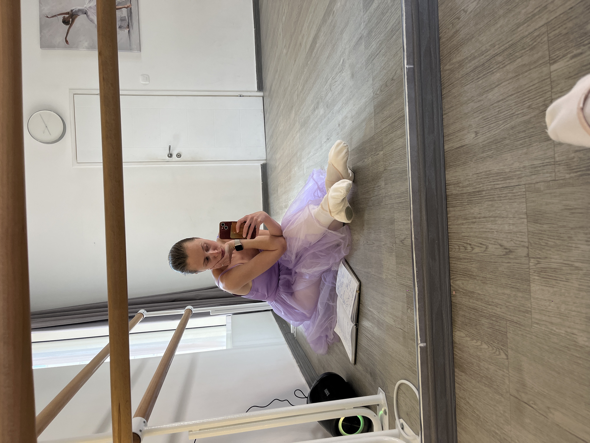 A young woman wearing a lavender leotard and tutu is seated on a light grey wood floor, facing and taking a selfie in a mirror in what appears to be a dance studio or home studio. She is wearing ballet shoes and has a book open next to her. In the background, there is a ballet barre, a white door, a clock, and a painting of a ballet dancer. The environment suggests an indoor space, and based on the style of the room and available information, this could potentially be in Budva, Montenegro, or more likely, a replica of one.