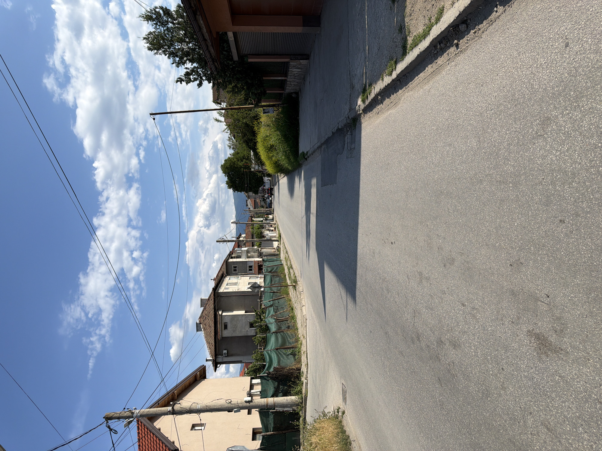 The image shows a street scene in Plovdiv, Bulgaria. The street is paved with asphalt and has a sidewalk on the right side. There are houses on both sides of the street, with a green fence along the left side. Overhead power lines are visible. The sky is blue with some clouds. In the distance, there are more houses and trees. The overall impression is of a residential street in a suburban area.
