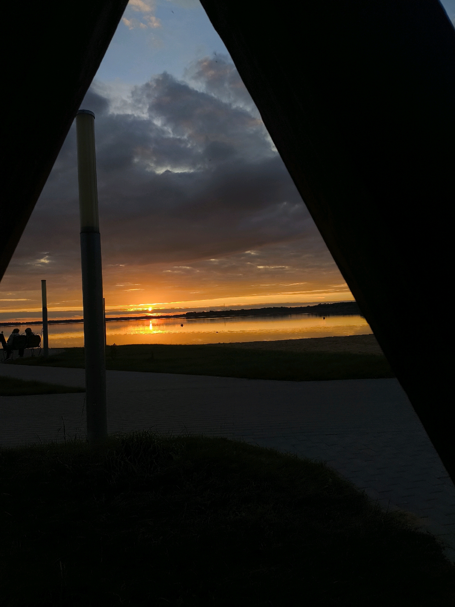 The image depicts a sunset over a body of water, possibly Lake Myadel, in Myadzyel, Belarus. The sun is low on the horizon, casting a vibrant orange and yellow glow across the sky and reflecting on the calm water. Dark clouds are present in the upper part of the sky, contrasting with the bright sunset. In the foreground, a dark, triangular shape frames the scene, suggesting the image is taken from within a structure or an opening. On the left side of the image, a street lamp and a few silhouetted figures are visible sitting on a bench near the water. A paved walkway runs along the water's edge, bordered by a grassy area. The overall atmosphere is serene and tranquil, characteristic of a peaceful evening by the lake.
