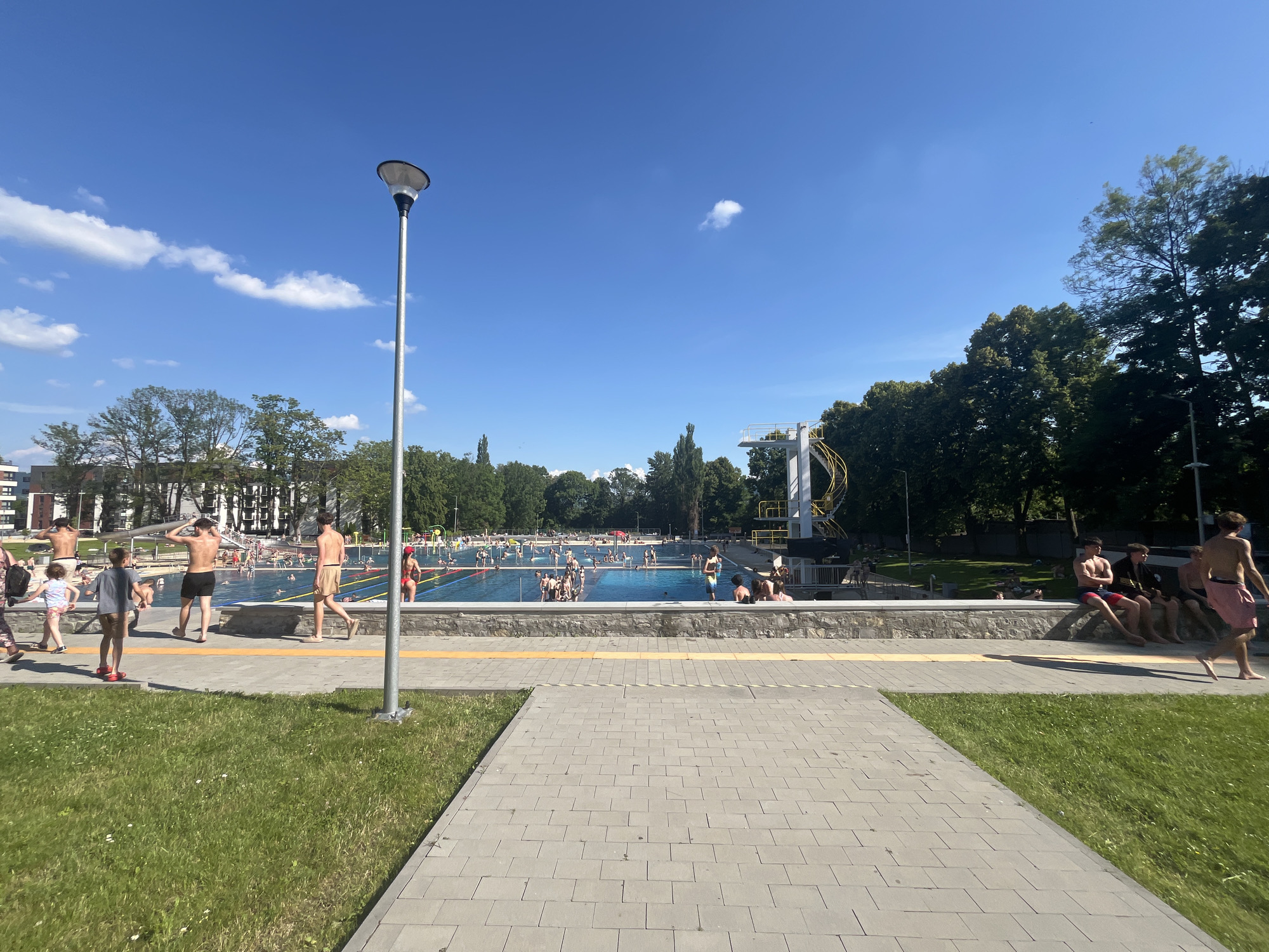 An outdoor swimming pool complex is depicted on a sunny day with a clear blue sky and scattered white clouds. The foreground shows a paved walkway and a grassy area. Beyond the walkway is a raised concrete wall with a stone facade, bordering a large, blue swimming pool filled with people. Some individuals are swimming, while others are standing or sitting around the pool's edge. A diving platform with yellow railings is visible on the right side of the pool. Trees and buildings form the background. Several people are present, including adults and children, wearing swimwear. A tall lamppost stands on the left side of the paved area.