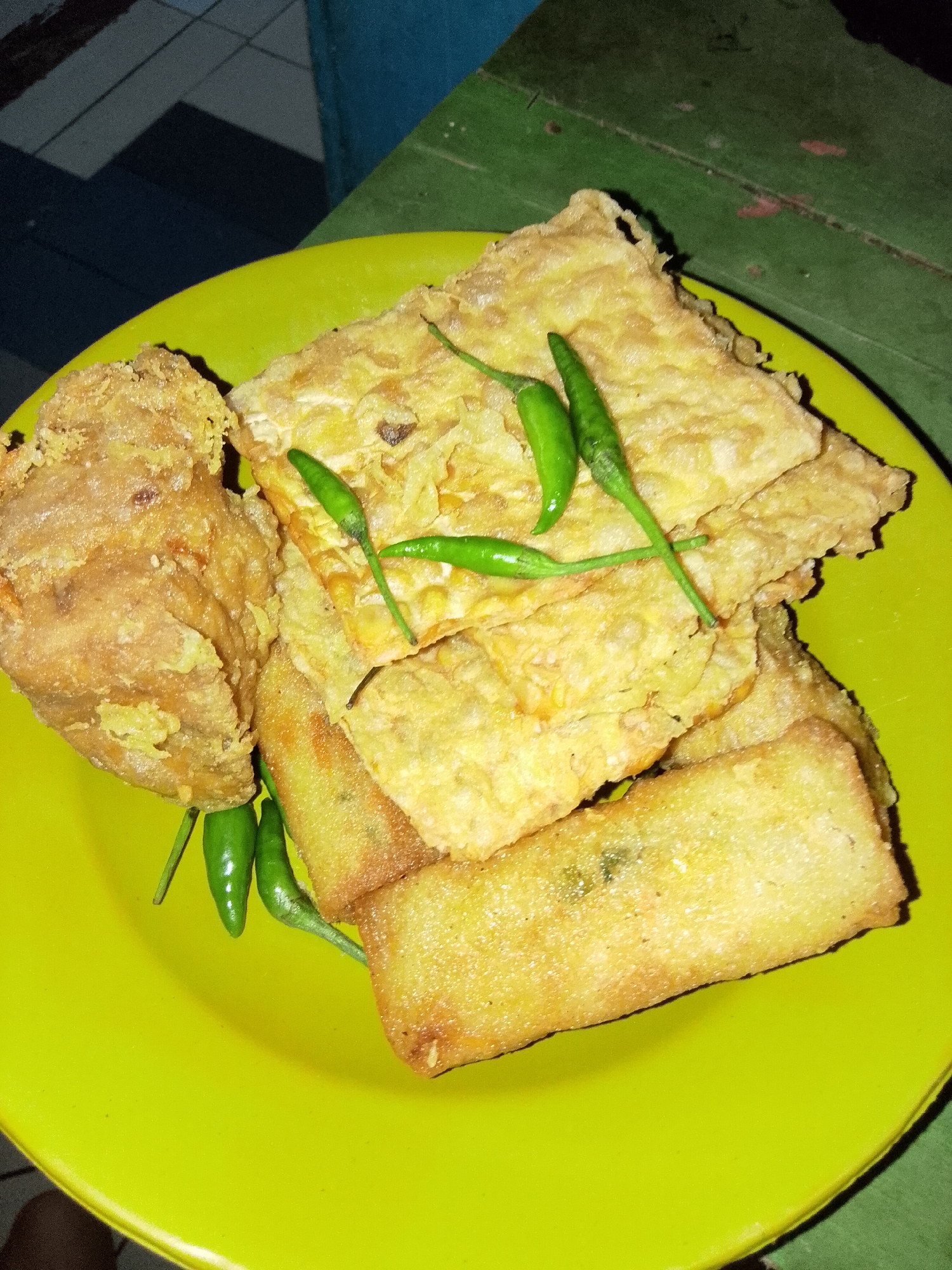 A plate of fried food, including what appears to be tempeh and possibly a piece of fried tofu, is presented on a bright yellow plate. Several small, green chilies are scattered on top of and around the fried items. The food is golden brown and appears crispy. The plate is positioned on a green wooden surface, possibly a table. In the background, a tiled floor and a blue wall are visible. The image is taken from a close-up, slightly overhead perspective.