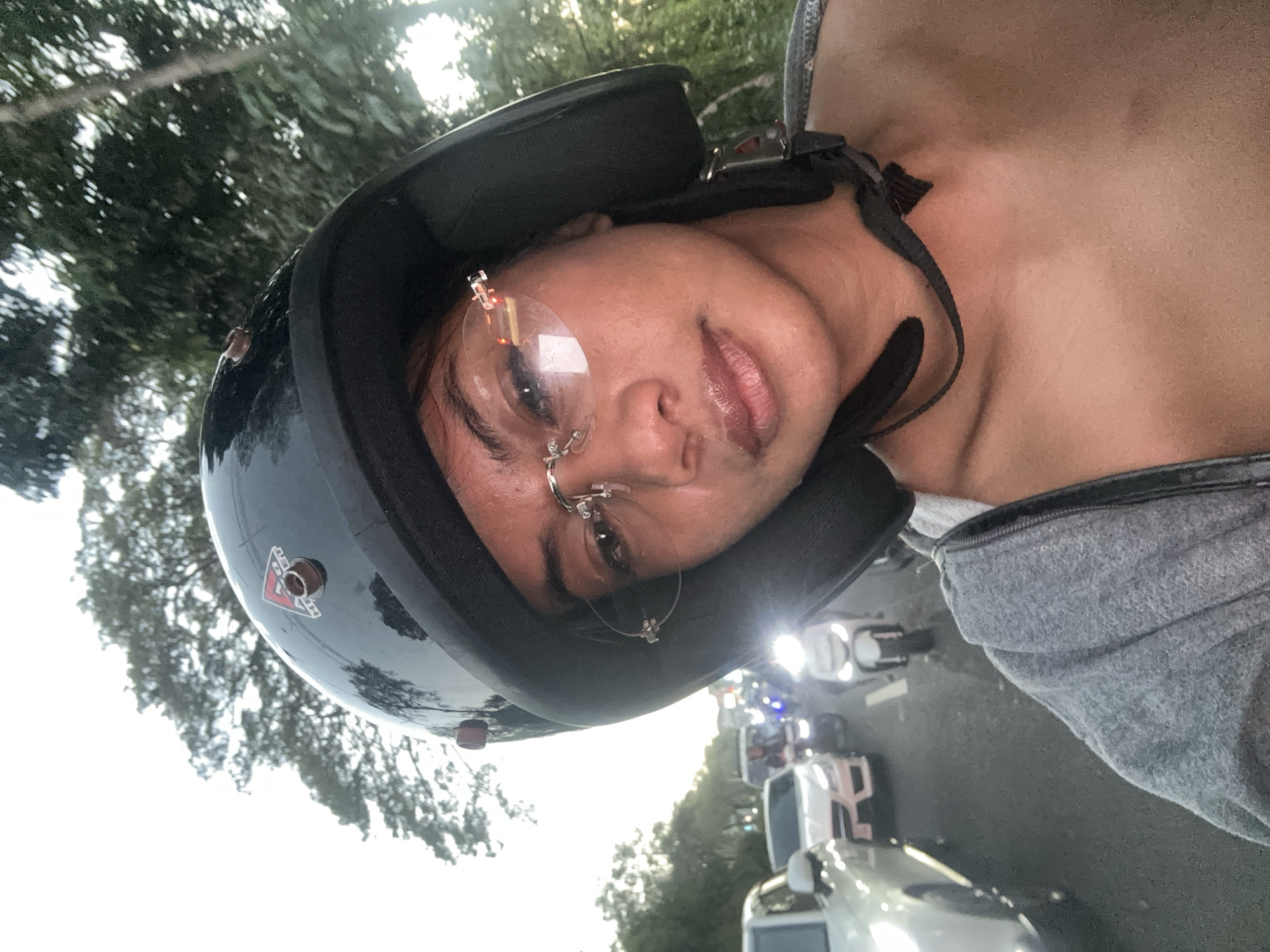 A close-up, low-angle selfie taken by a person wearing a black helmet and rimless glasses. The person is looking forward, with traffic visible behind them. The background includes trees and a cloudy sky, suggesting an outdoor setting in Denpasar, Indonesia, possibly during traffic congestion. The person is wearing a grey zip-up hoodie. The image appears to be taken from the perspective of someone on a motorcycle.