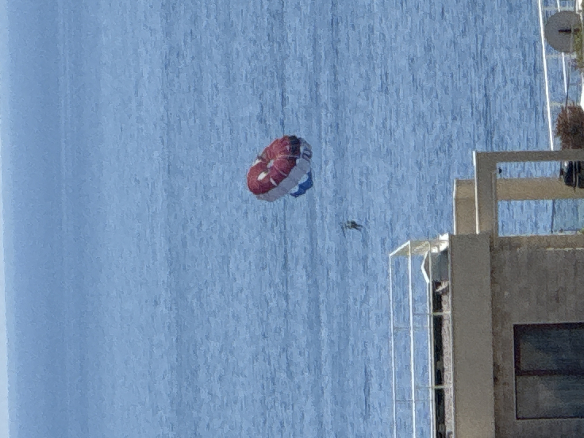 In Boreti, Montenegro, a parasailer is visible in the blue ocean, with a red and white parachute overhead. A person is suspended beneath the parachute. In the foreground, the top of a beige building with a railing and a window is visible. The building appears to be a hotel or apartment complex. The water is calm with small ripples, suggesting a clear and sunny day.