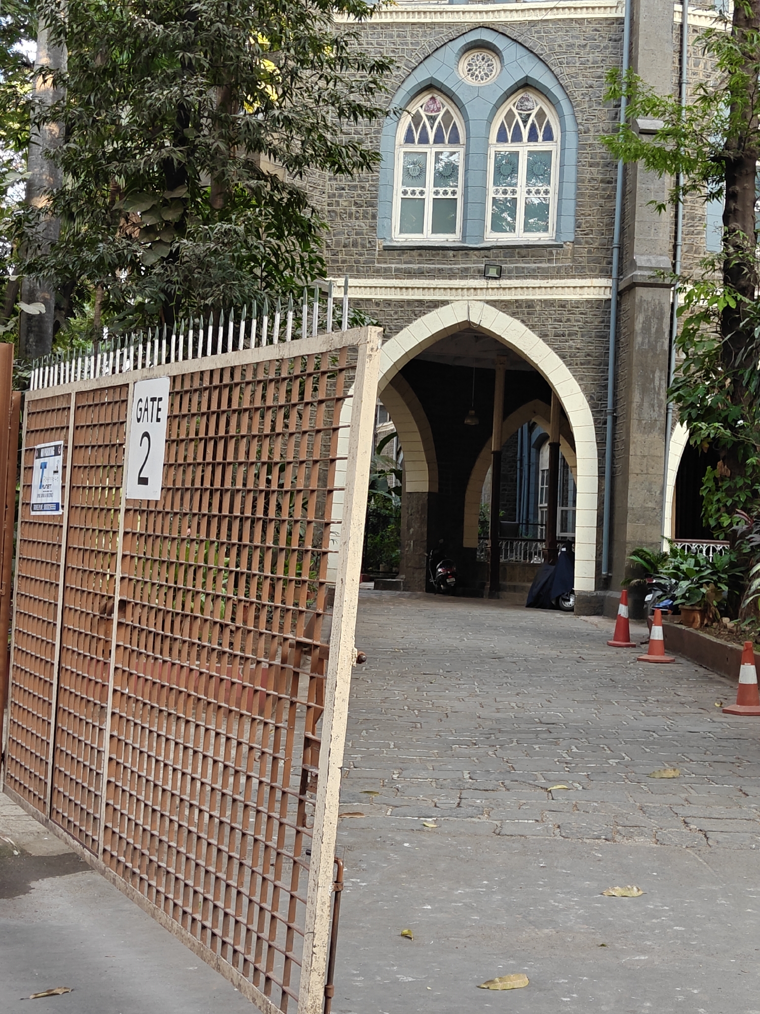 A view of a gate labeled "GATE 2" provides entry to the grounds of a stone building. The building has arched entrances and Gothic-style windows with decorative elements, suggesting it might be a church or a historical building. A series of traffic cones are visible inside the gated area. The paving is of cobblestones. Based on architectural style and the prevalence of such structures in Mumbai, India, and the presence of a sign in English, it is likely located in Mumbai.