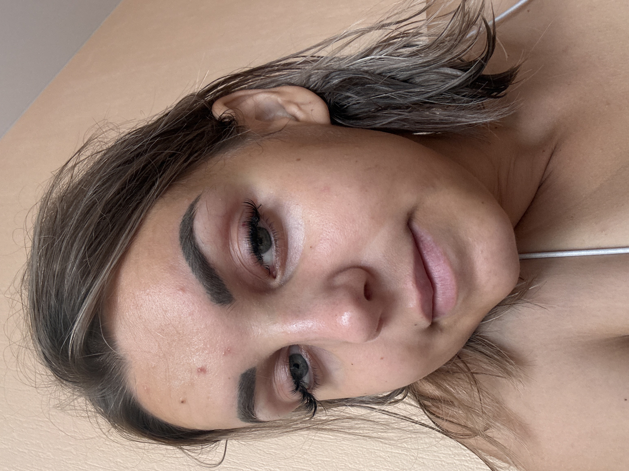 A young Caucasian woman with shoulder-length brown hair, styled in loose waves, is captured in a close-up, slightly angled, selfie-style portrait. Her complexion is fair with visible pores and several small blemishes, particularly on her forehead and cheek. She has prominent eyebrows, dark eyelashes, and appears to be wearing minimal makeup. Her eyes, a muted green, are looking slightly off-camera. Her lips are slightly parted, revealing a natural pink hue. The background is a plain, textured wall in a pale peachy-beige color. The lighting is soft, suggesting an indoor setting. The woman is wearing a white strap, possibly from a tank top or dress, visible at the bottom of the frame. The image does not provide any information that would allow for determining the location within Kholmsk, Russia.