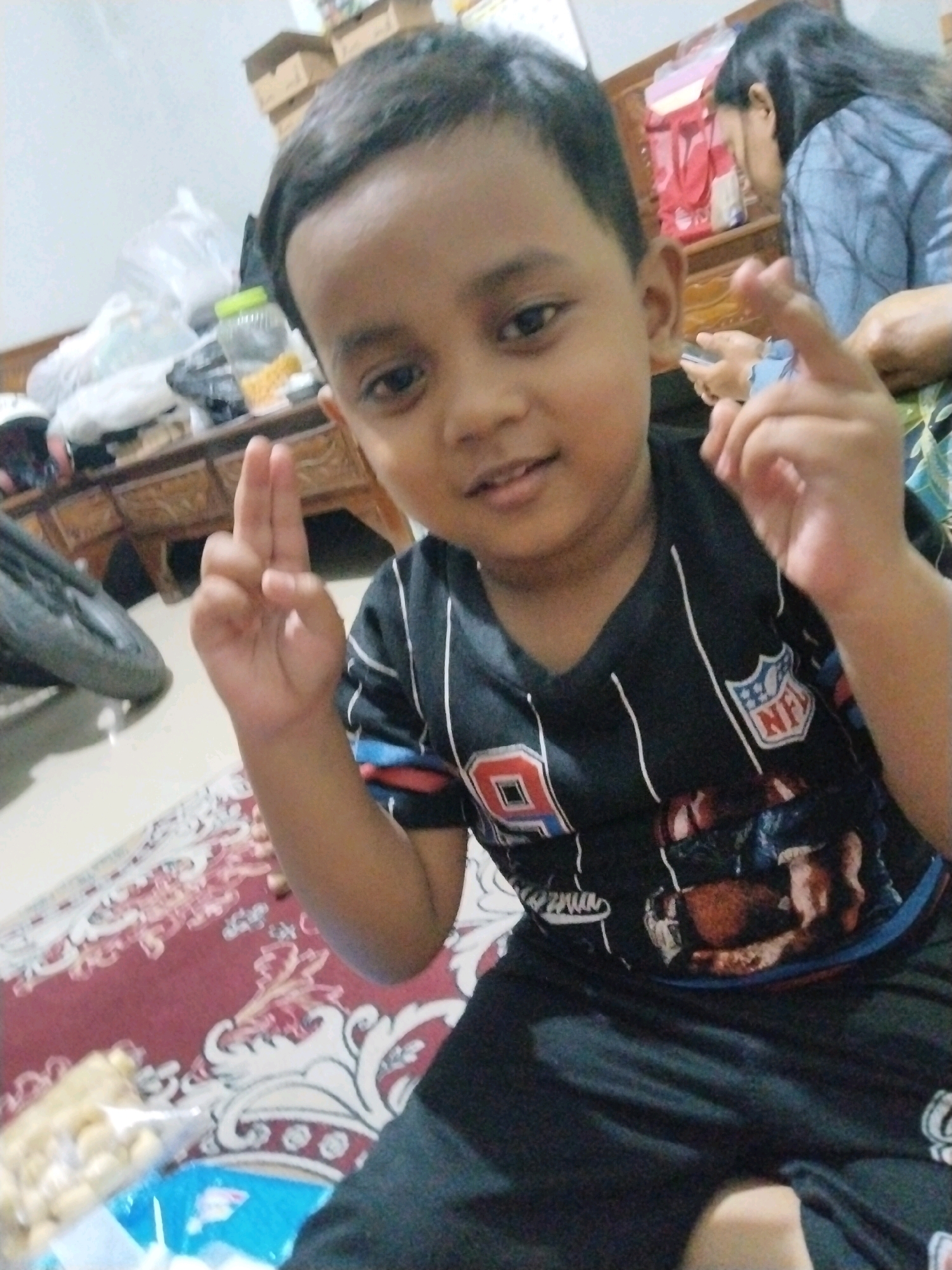 A young boy, possibly of Indonesian descent and appearing to be around 4-6 years old, is the central subject of this indoor photograph. He is captured from the chest up, facing the camera with a neutral expression and a slight smile. He has dark hair, dark eyes, and brown skin. He is wearing a black t-shirt with white stripes on the sleeves and a graphic print on the front, which includes the NFL logo and what appears to be an image of football players. His right hand is raised, with his index and middle fingers extended upwards, forming a "V" shape. The background is somewhat cluttered, suggesting a residential environment. To the left, a table is visible with various items, including what looks like a pile of plastic bags and containers with yellow objects inside, possibly snacks. A portion of a red and white patterned rug is in the lower part of the image, and a black object, possibly a motorcycle wheel or tire, is also partially visible. In the upper right background, a woman with long dark hair is partially visible, looking down and holding a phone. The setting appears to be a home in Pepelegi, Indonesia.