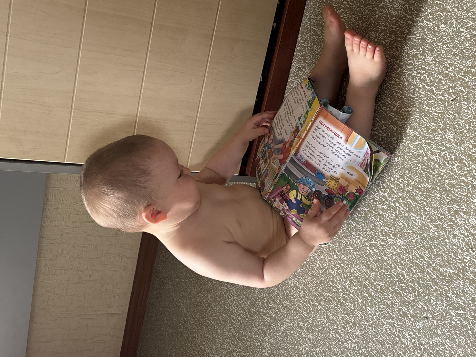 A light-skinned toddler, wearing no clothes, sits on a patterned carpet, leaning against a light wood cabinet with horizontal grooves. The toddler is looking down at an open book with colorful illustrations and text. The book is spread open in front of the child, with the left page depicting a scene with a child on a bicycle and the right page showing text and some illustrations, including what appears to be a bird. The background wall has a textured, light brown wallpaper. The scene is indoors, likely in a residential setting in Naberezhnye Chelny, Russia, based on the provided context. The lighting suggests daytime.