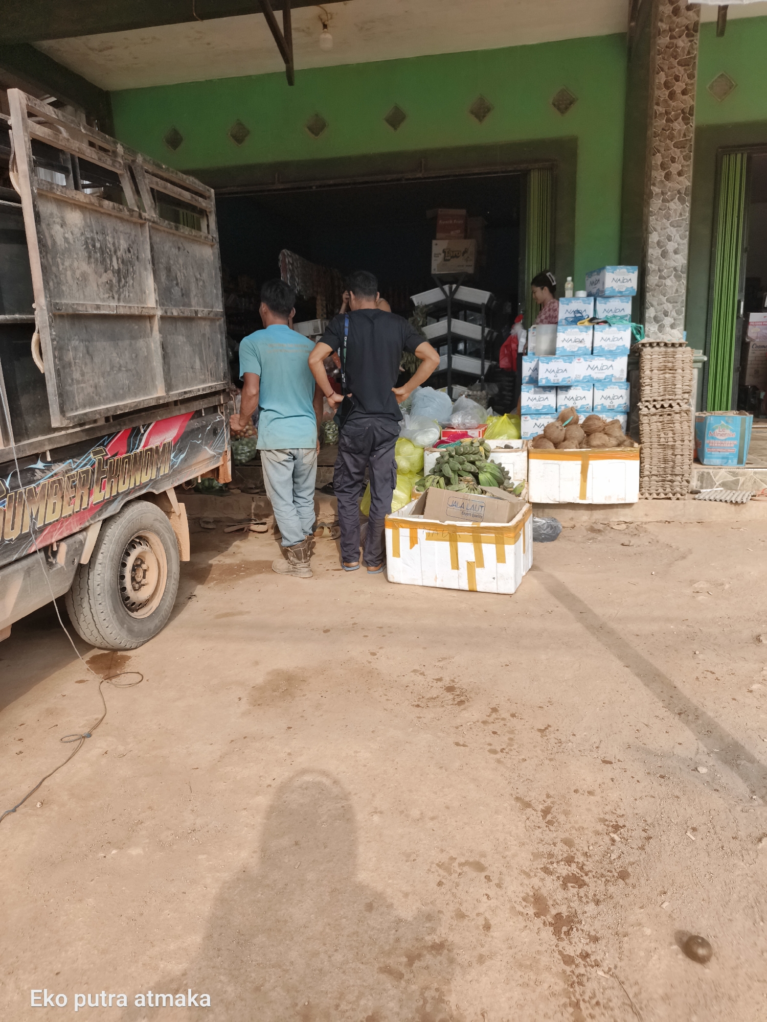 A truck with a loaded bed is parked in front of an open-air market stall in what appears to be Selimantan Jaya, Indonesia. Two men, one in a blue t-shirt and the other in a black t-shirt, stand near the truck, appearing to be unloading or organizing goods. The market stall is stocked with various items including crates of bottled water, egg cartons, and piles of bananas and coconuts. A woman is visible in the background inside the stall. The ground is dusty and appears to be a dirt road or unpaved area. The overall scene suggests a commercial transaction or delivery of goods.