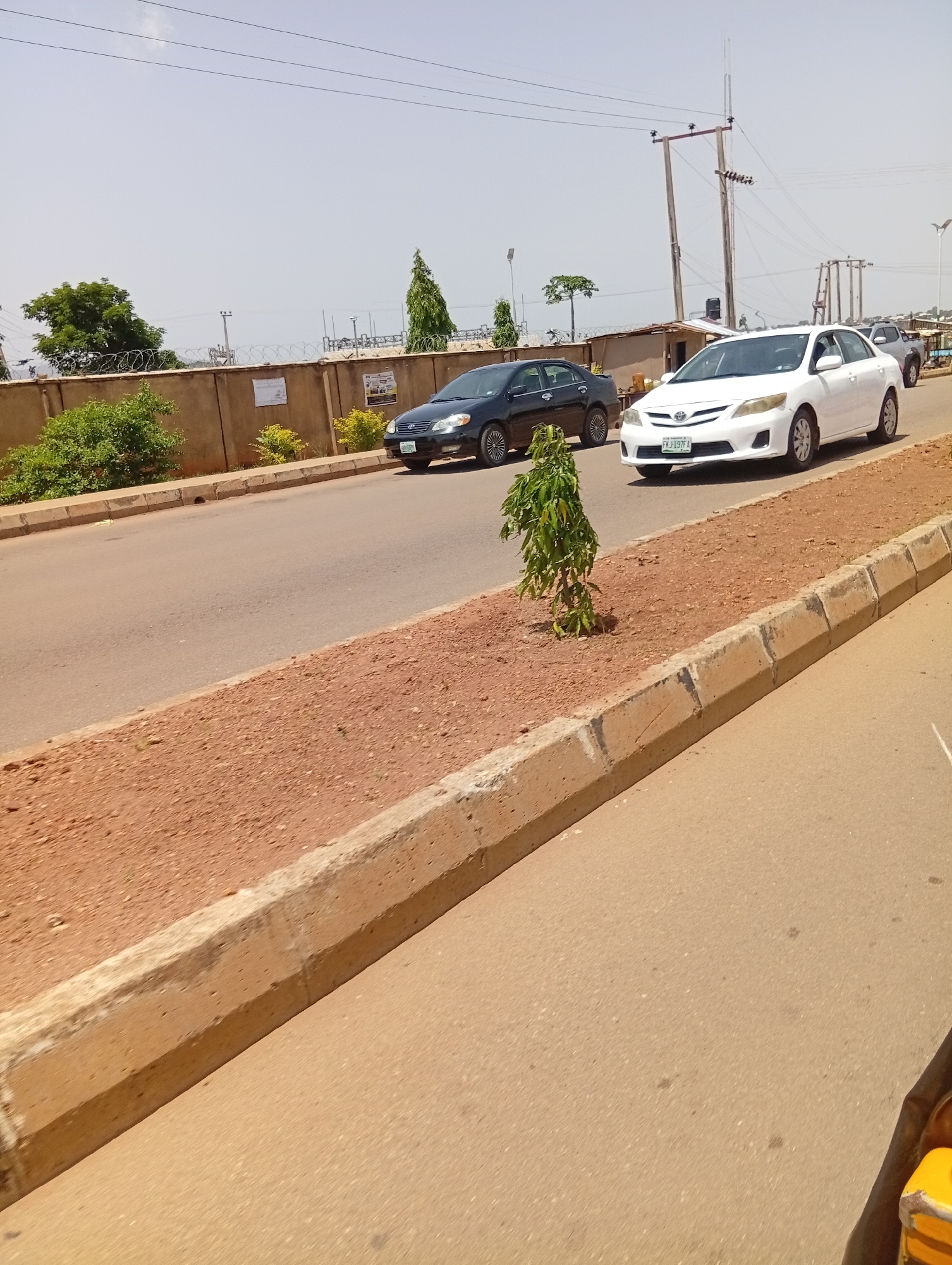 The image shows a street scene in Jos, Nigeria, on a bright, sunny day. The foreground is dominated by a paved road and a concrete curb, with a strip of reddish-brown soil containing a small, leafy tree.
In the middle ground, two cars are driving on the road. The car closest to the viewer is white, and the one behind it is black. The white car has a license plate that appears to read "FKJ 197 FA". Beyond the road, a concrete wall with barbed wire runs along the side of the street, separating it from an area with some vegetation and a few structures, including what looks like a small shed. Utility poles and power lines are visible in the background, suggesting an urban or semi-urban setting. The sky is clear and pale, typical of a hot day.
The scene depicts everyday traffic and infrastructure in Jos, Nigeria. The bright sunlight indicates it is likely daytime, possibly late morning or early afternoon. There are no people clearly visible in the image, and the overall impression is one of ordinary daily activity on a road.