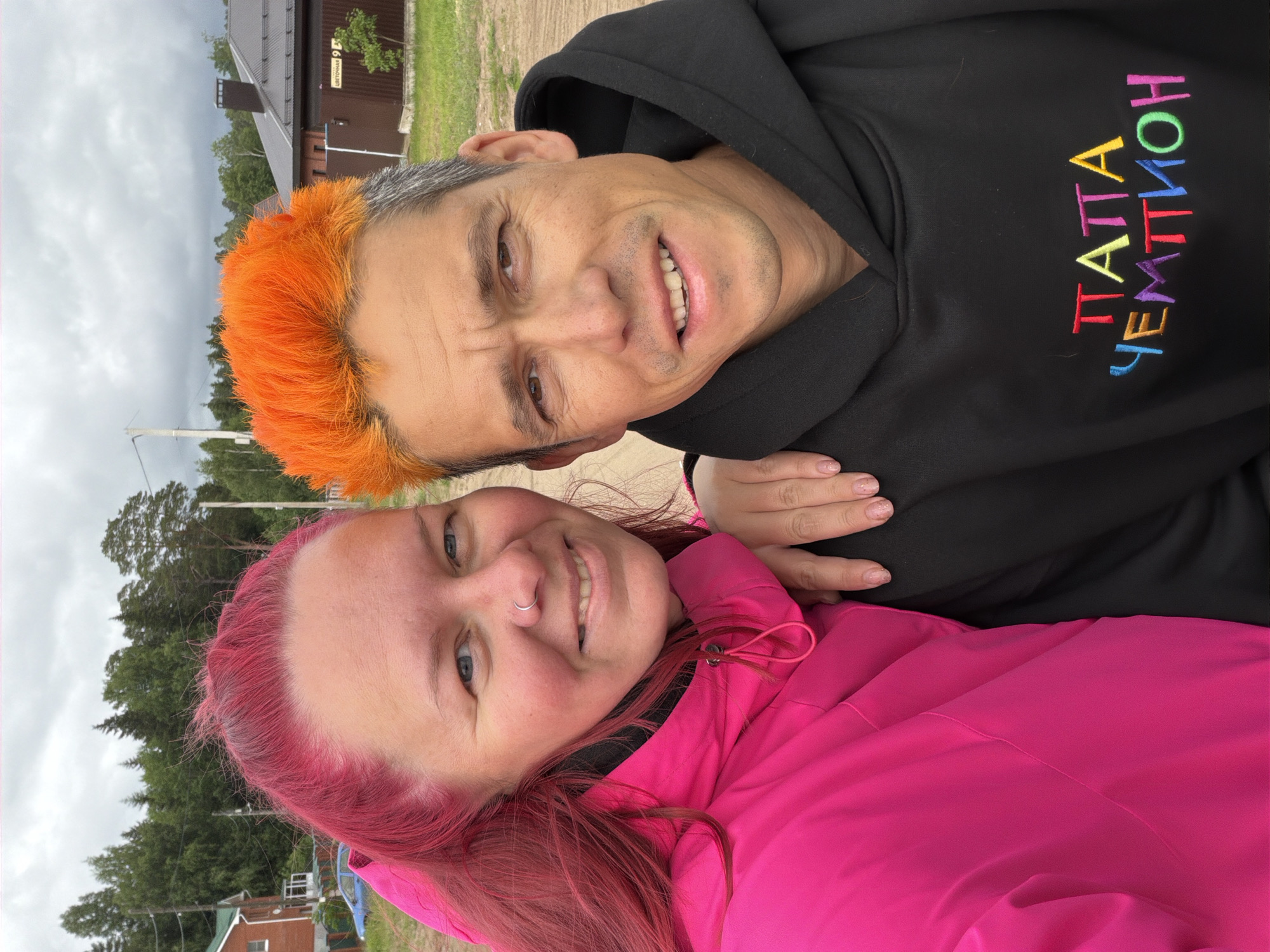 This detailed description outlines a selfie-style photograph featuring two adult individuals, a female and a male, in an outdoor setting in what appears to be a residential area in Chaikovsky, Russia.
The female, positioned on the left, has bright fuchsia-pink hair, blue eyes, and fair skin. She is smiling, revealing her teeth. A silver hoop nose ring is visible on her left nostril. She wears a vibrant pink hooded jacket, possibly with a black layer underneath. Her right hand, adorned with light-colored, possibly glittery, nail polish, rests on the male's left shoulder. She appears to be in her 30s to 40s.
The male, on the right, has distinctively dyed orange hair on top, contrasting with natural gray hair visible on the sides and temples. He has brown eyes, a tan complexion, and is smiling subtly. He wears a black hooded sweatshirt with colorful embroidered text on the chest. The text, in Cyrillic, reads "ПАПА ЧЕМПИОН" (PAPA CHEMPION), translating to "DADDY CHAMPION" in multi-colored block letters (red, orange, yellow, green, blue, purple, pink). He also appears to be in his 30s to 40s.
The background indicates an overcast day with a cloudy sky. The immediate foreground shows a dirt or gravel surface. Behind the individuals, green grass is visible on the right. In the mid-ground and background, various trees, including conifers, are present. Buildings include a two-story red brick house with a green roof and white window frames on the left, near which a light blue car is partially visible. On the right, a darker-colored house or building with a dark roof and chimney is visible. A white street sign on a pole near this building clearly displays "УЛИЦА ЦВЕТОЧНАЯ 9" (ULITSA TSVETOCHNAYA 9), confirming a Russian location.