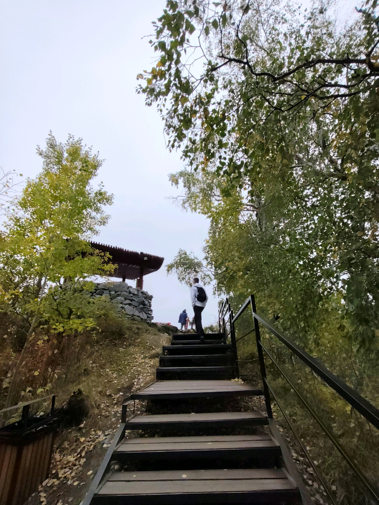 The image shows an outdoor staircase leading up to a stone structure topped with a wooden pavilion in Nikola, Russia. A person wearing a light-colored hoodie and a backpack is walking up the stairs, facing away from the camera. Another person in darker clothing is visible further up the stairs, and a third person is near the pavilion. The staircase has metal railings on the right side and is constructed with dark wooden steps. The surrounding environment is characterized by trees with green and yellowing leaves, indicating it is likely autumn. Fallen leaves are scattered on the ground and along the path next to the stairs. The sky is overcast. The stone structure appears to be a base for the pavilion, built with irregularly shaped rocks.