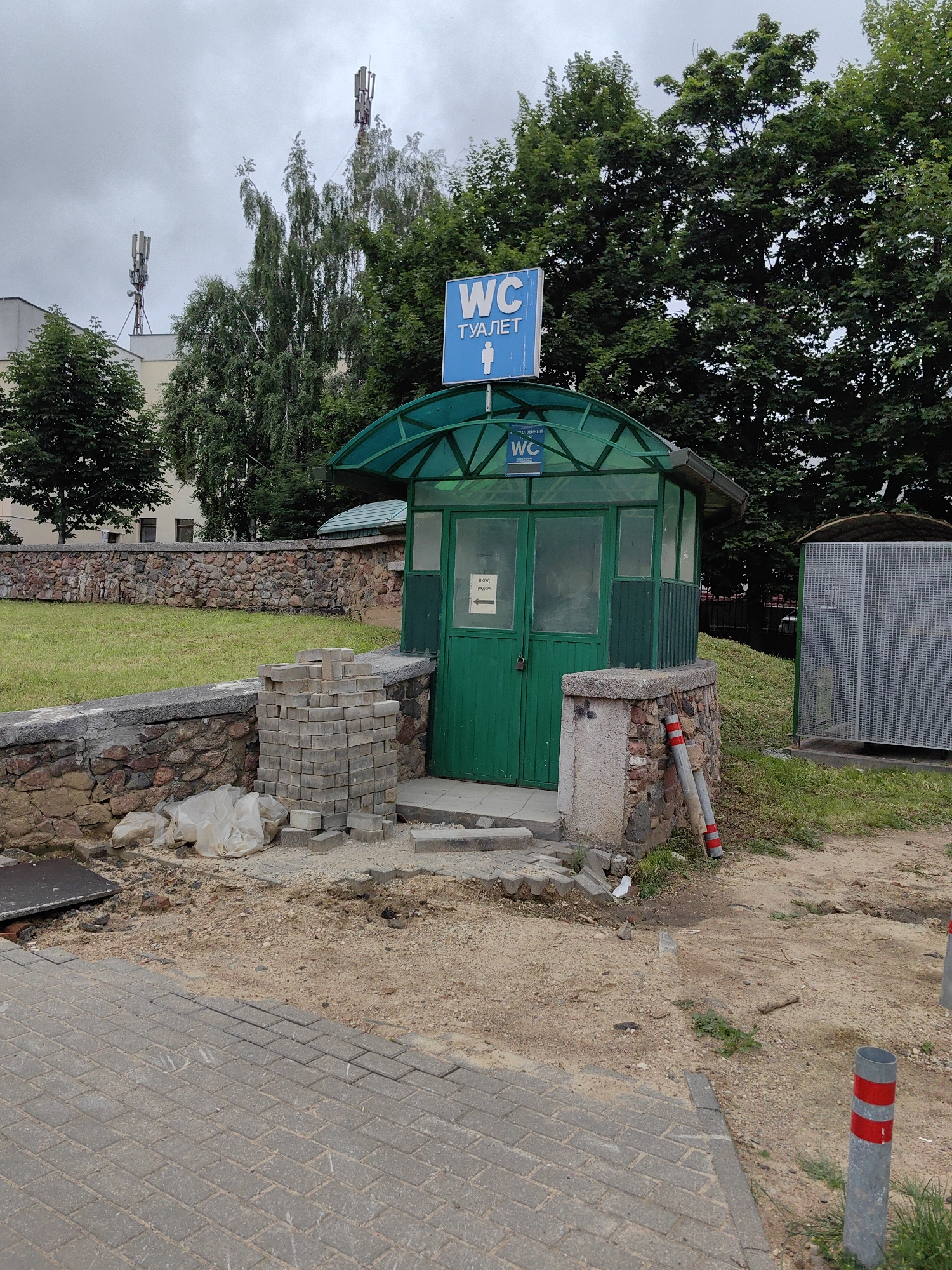 The image depicts a public toilet facility, likely in an outdoor urban setting. The toilet structure itself is green with a transparent green canopy. A blue sign with white lettering, indicating "WC" and "ТУАЛЕТ" (Russian for toilet), is visible above the entrance, along with a pictogram of a person. Another smaller "WC" sign is affixed to the canopy. The area surrounding the toilet appears to be under construction or undergoing maintenance. Piles of gray paving stones are stacked near the entrance, and loose stones and sand are present on the ground. White plastic sheeting, possibly covering materials, is also visible.
To the left of the toilet, a low stone wall topped with a concrete coping separates the paved walkway from a grassy area. Behind the wall, a building with visible windows can be seen, partially obscured by trees. In the background, tall trees dominate the scene. Two cellular towers are prominent against the cloudy sky, situated atop buildings.
To the right of the toilet, a large, metallic, mesh-covered structure stands. Two cylindrical bollards with red and white reflective bands are positioned in the foreground, marking a boundary. The ground in the immediate foreground is paved with gray interlocking bricks, leading to a sandy, uneven surface where the construction materials are located.
The overall environment suggests a public space in a city, possibly a park or a street-side area. The presence of construction materials indicates ongoing work. The cloudy sky suggests overcast weather. This scene is located in Minsk, Belarus, as indicated by the context.