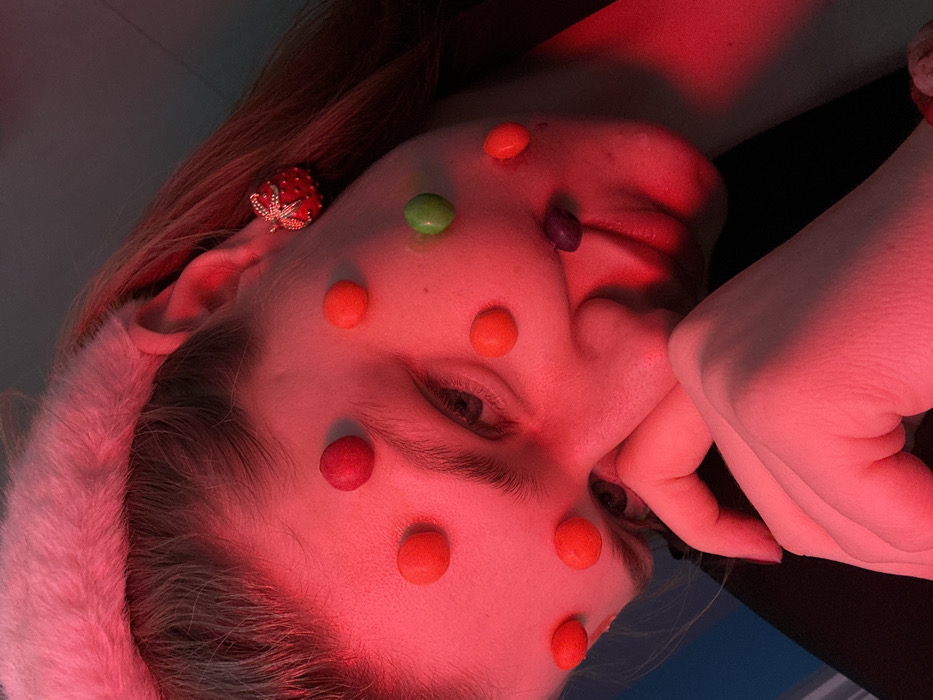 A young individual, likely female, with fair skin and light brown hair, is depicted in an indoor setting under intense red lighting. She wears a pink, fluffy headband or ear muffs and a dark garment. A red, textured earring resembling a strawberry with a small bow is present on her right earlobe. Her face is adorned with numerous small, round, candy-like objects: eleven red objects are distributed across her forehead, temples, and cheeks, one green object is on her right cheek, and one purple object rests on her chin. Her right hand, with fingers curled, is positioned near her chin. Her eyes are partially visible, looking directly forward. The background is indistinct, showing faint dark blue tones on the upper left. The specific location within the city of Kazan, Russia, cannot be determined from the visual information present in the image.