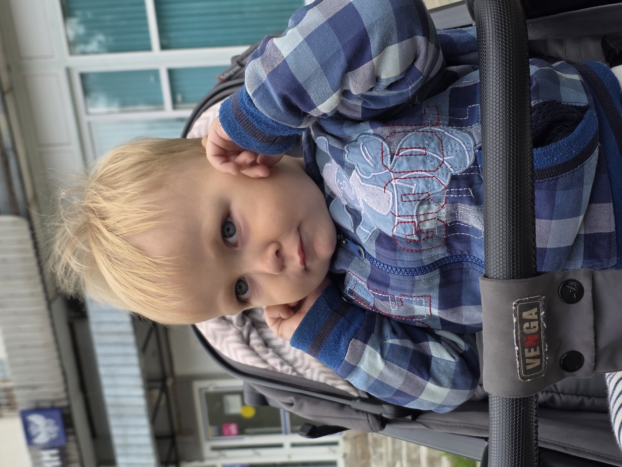 A blond-haired, blue-eyed toddler sits in a stroller. The toddler is wearing a blue plaid jacket with embroidered light blue lettering on the chest. The toddler's hands are raised to their ears, and their expression is thoughtful. The stroller has a grey fabric liner with a zigzag pattern and a black handle with a grey fabric cover that has the brand name "VEGA" visible. In the background, a building with a blue sign depicting a bird emblem is visible, consistent with a Russian Post office, suggesting the location is in the city of Chaikovsky, Russia. The overall environment appears to be outdoors, possibly on a street or sidewalk, with a building facade and windows in the background.